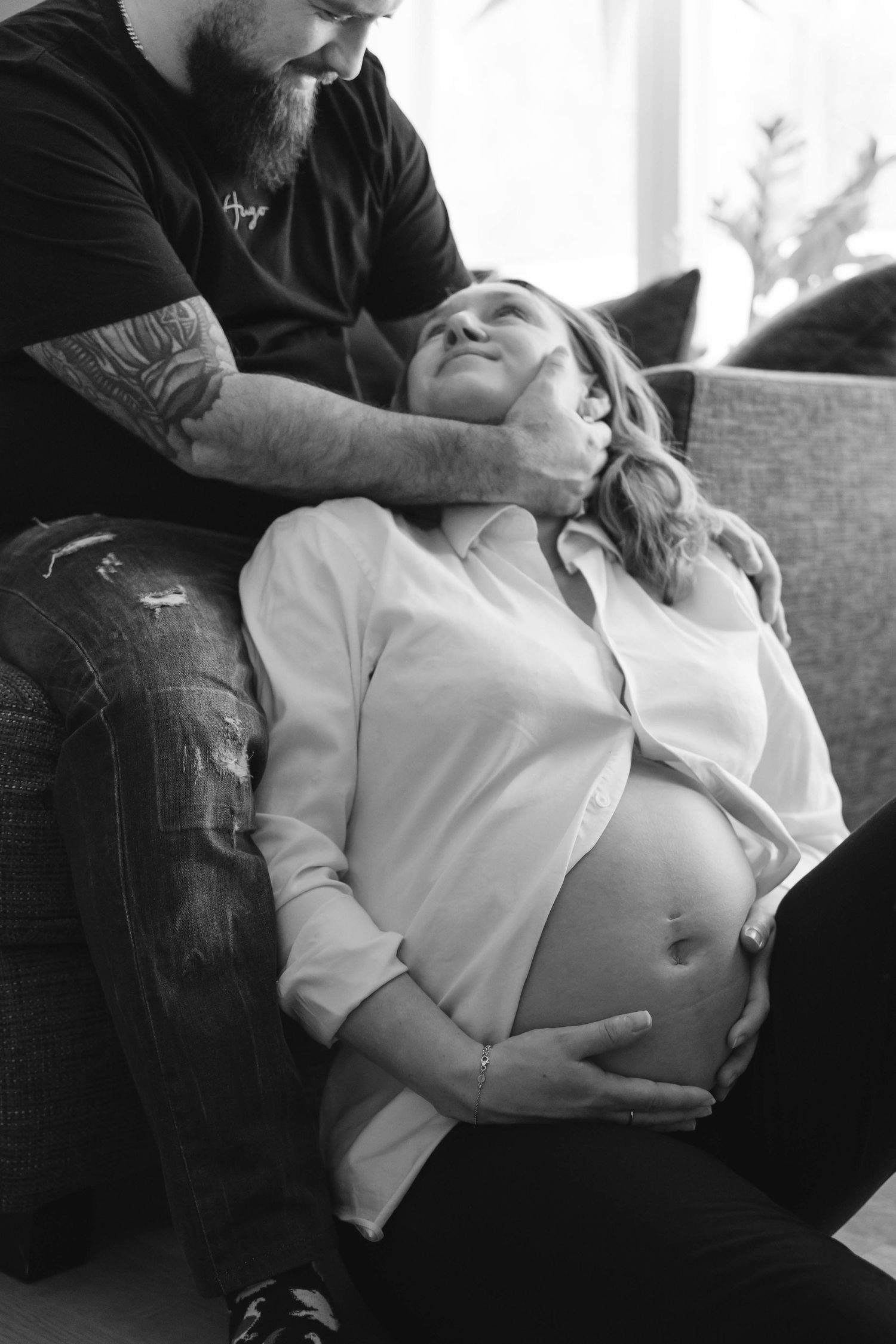 A black and white intimate maternity photo showing a couple relaxing together on a couch with exposed pregnant belly.