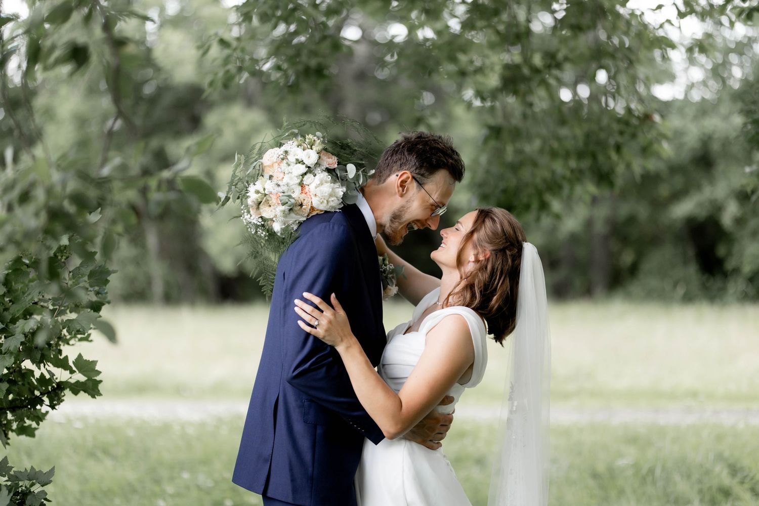 A bride and groom share a romantic kiss outdoors, surrounded by lush green trees on their wedding day.