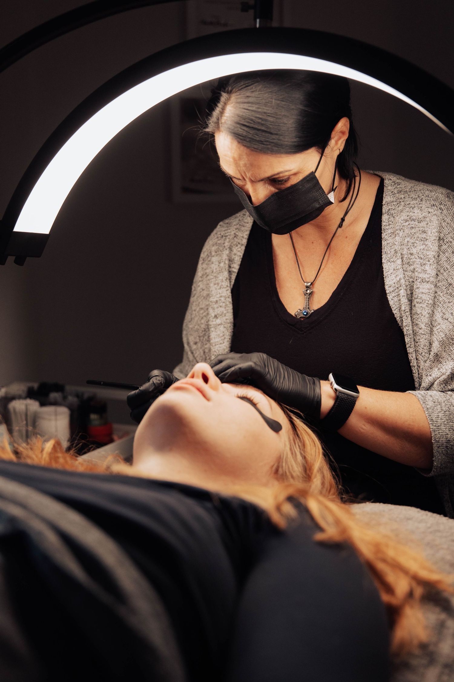 Professional beautician wearing protective mask and gloves performs an eyebrow procedure under bright LED light.