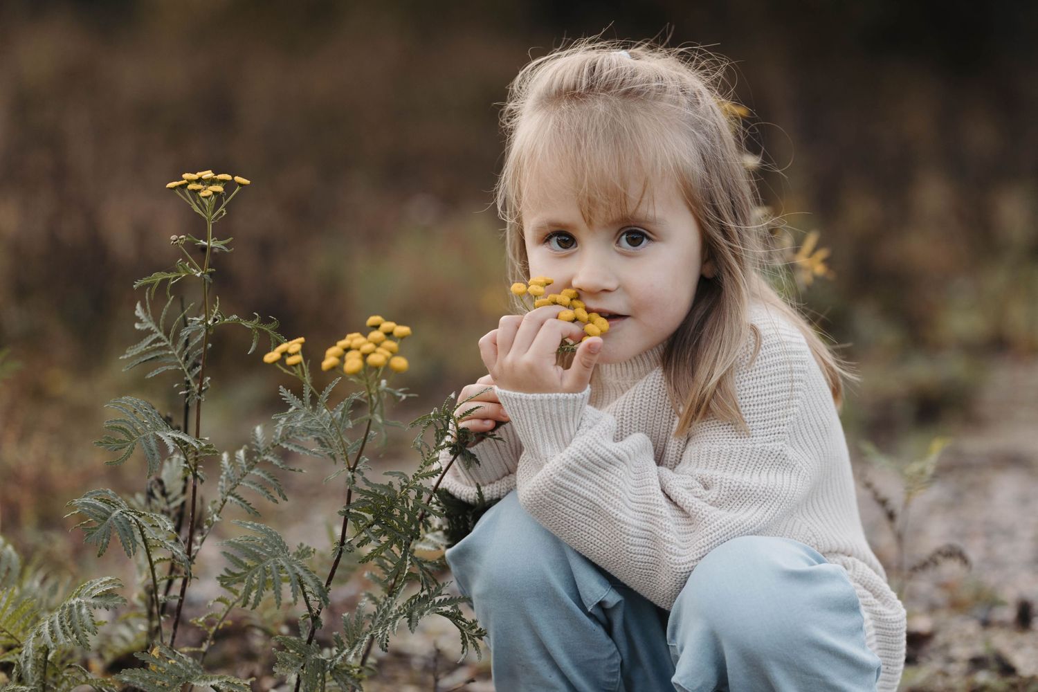A young child with blonde hair holds a yellow wildflower while sitting outdoors in a natural setting.