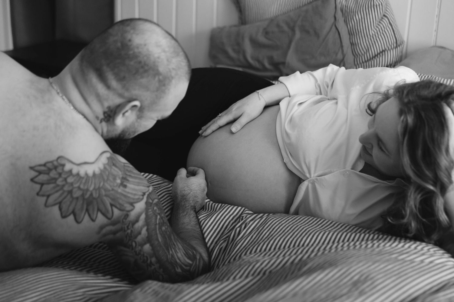 Black and white photo of a tattooed artist working on a tattoo design on someone laying on a bed.