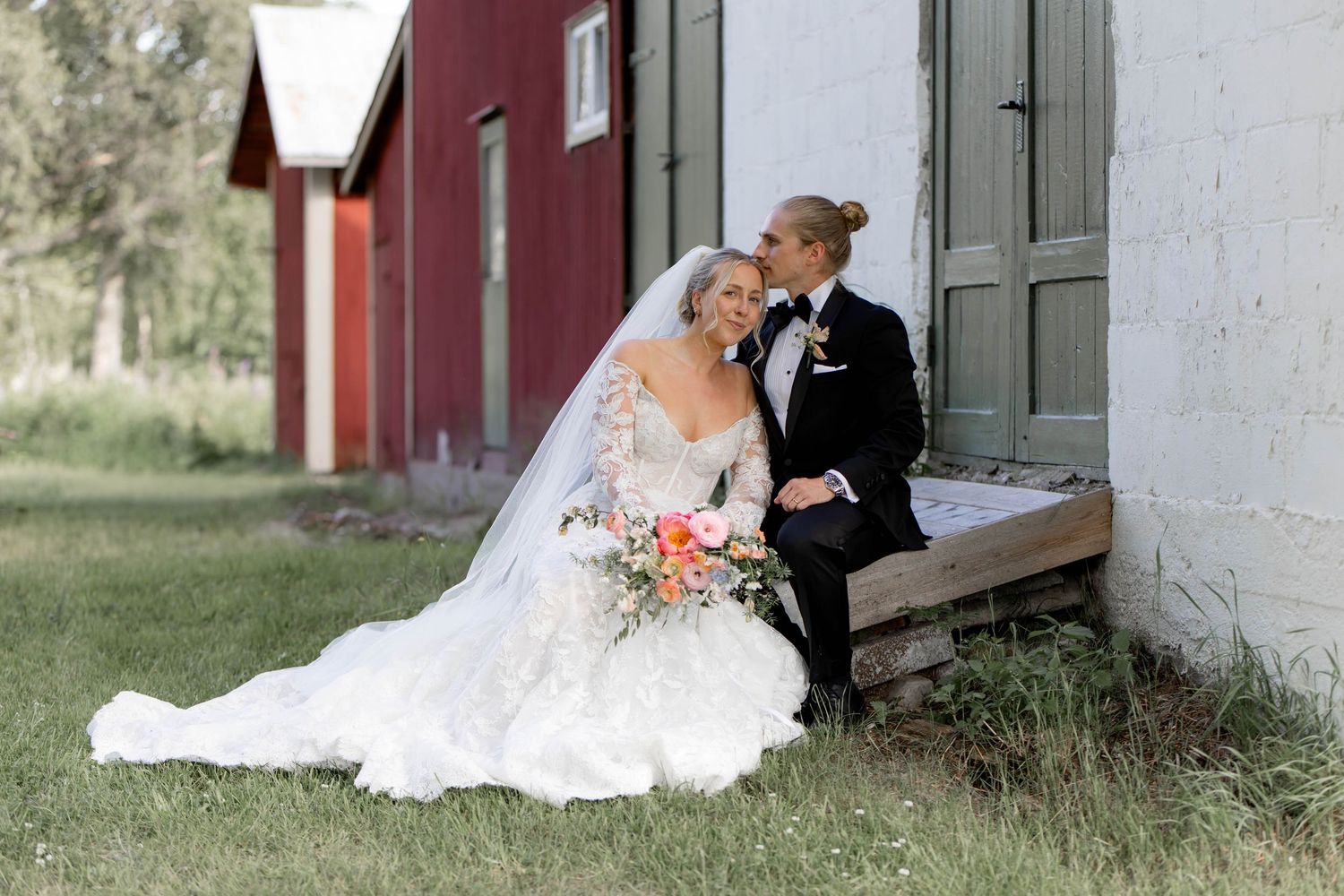 Bride in lace gown and groom in tuxedo share a kiss beside a rustic red barn on their wedding day.