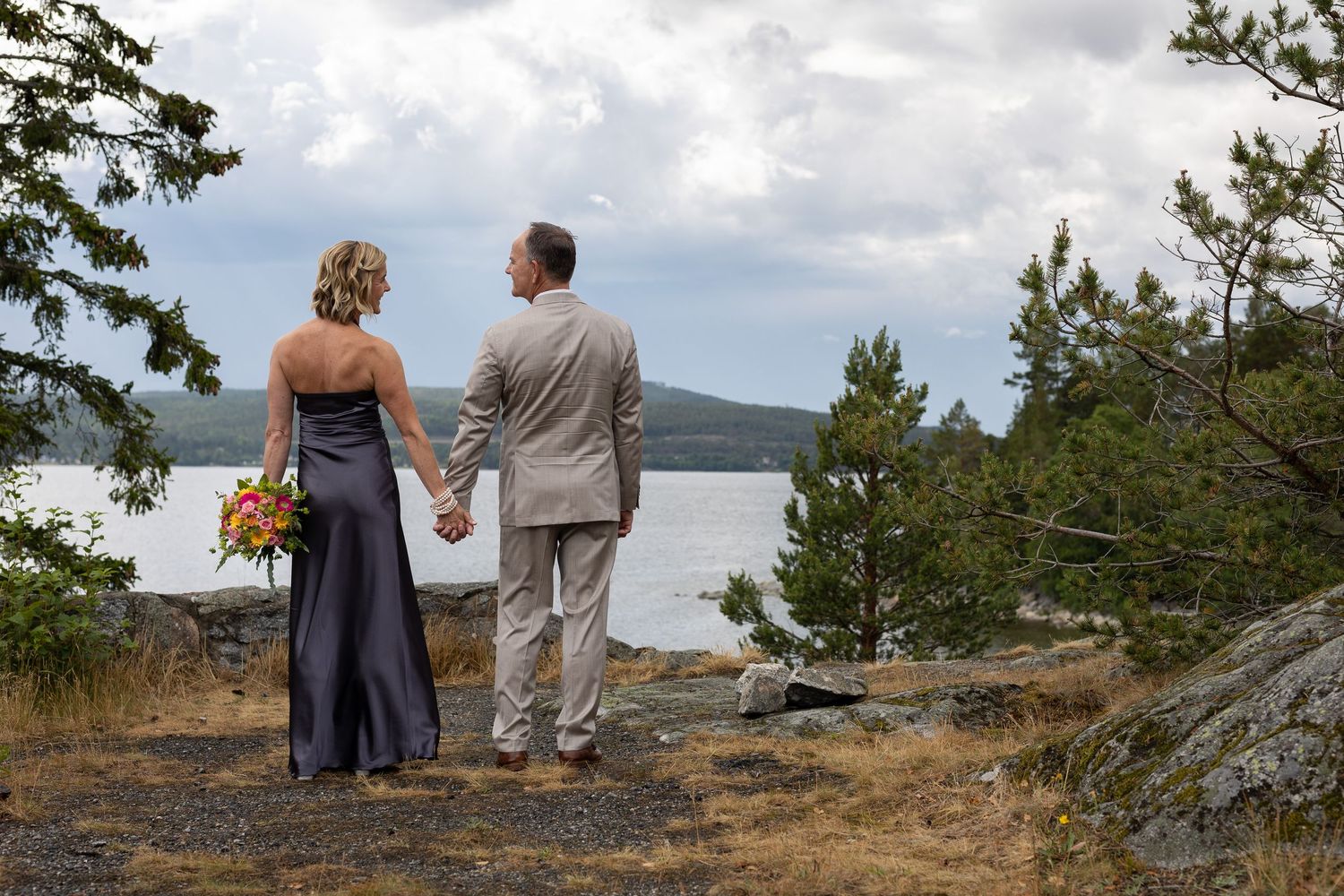 A couple sits together on a rocky coastal cliff overlooking the ocean on a sunny afternoon.