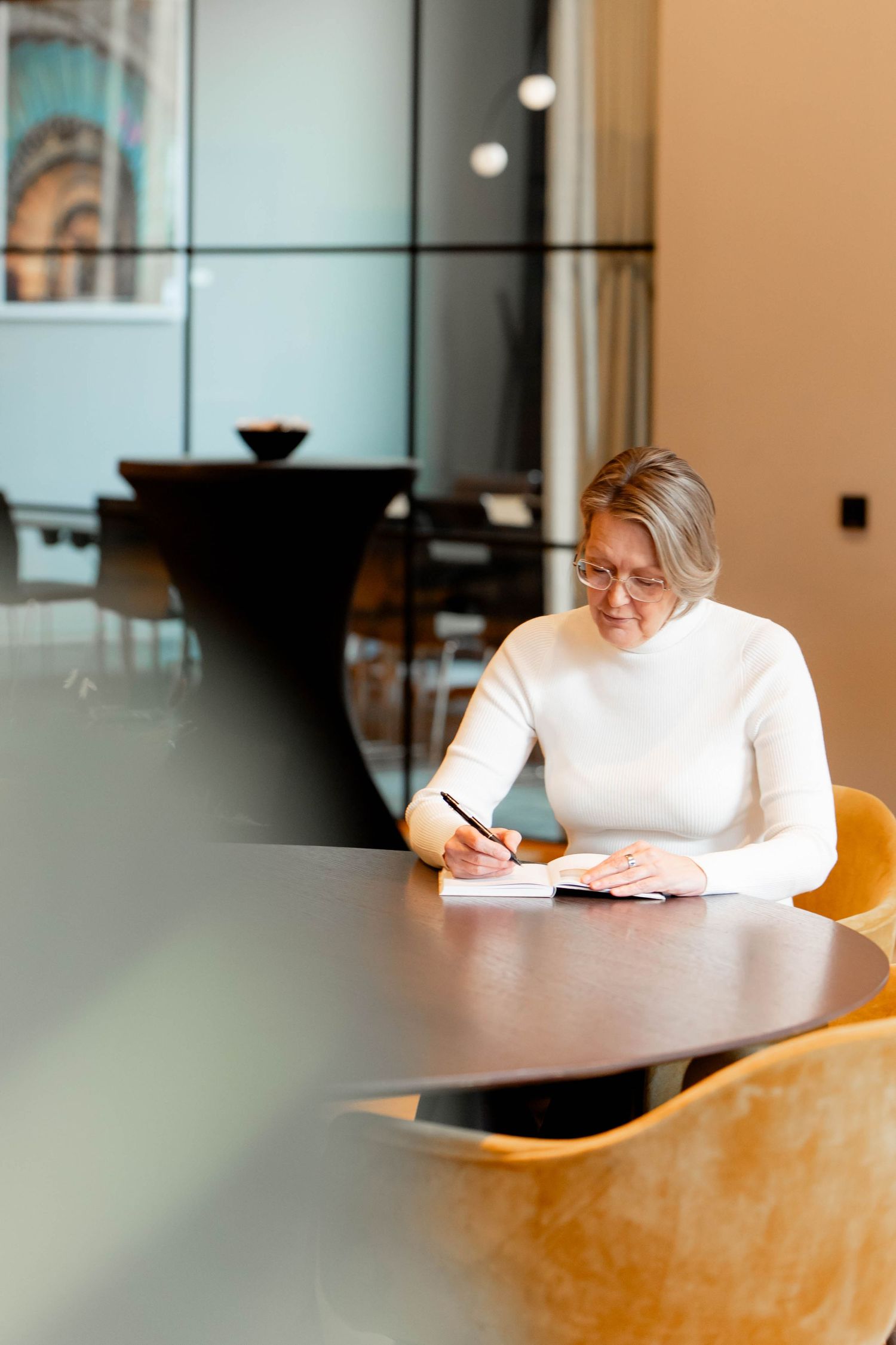 Person in white sweater writes in notebook while sitting at wooden table in bright modern office space.