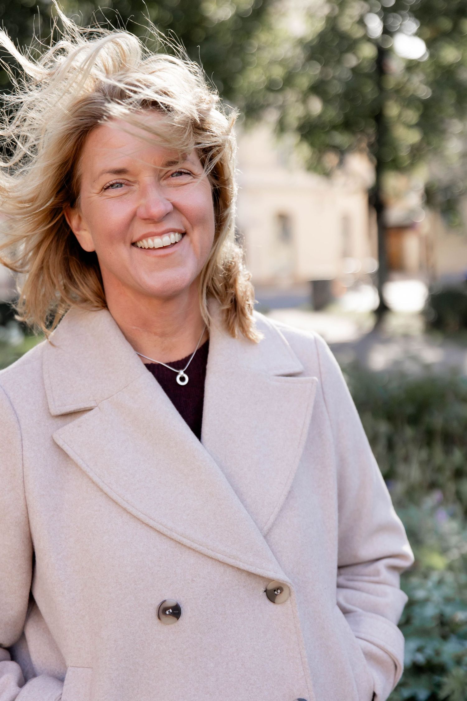 A person wearing a beige peacoat smiles outdoors on a sunny day with trees in the background.