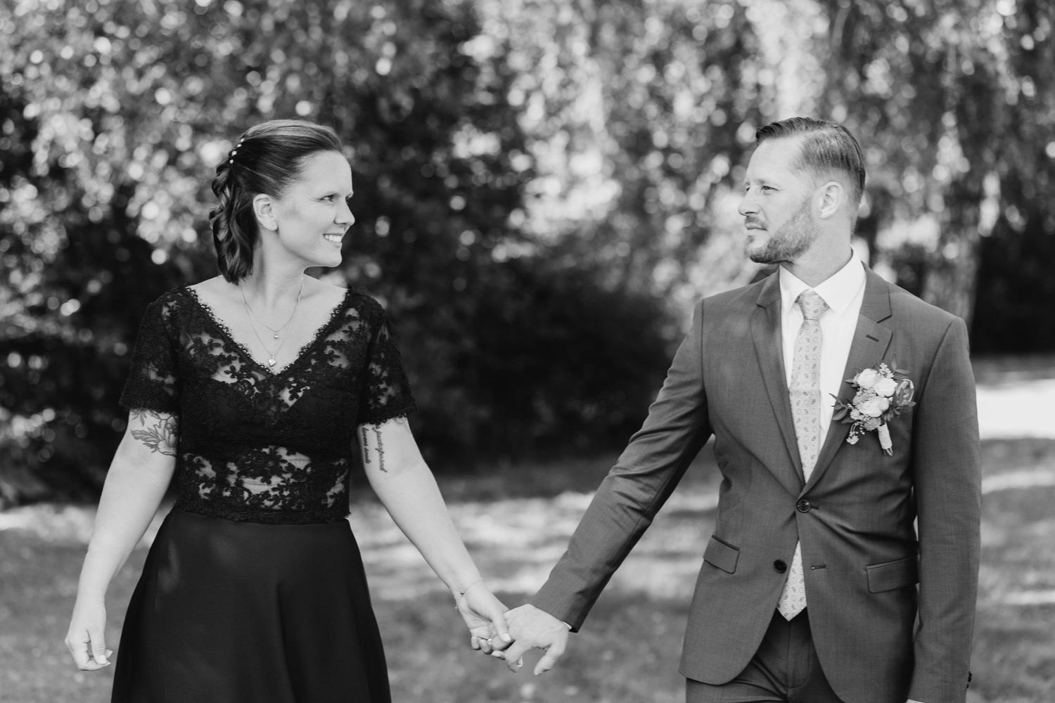 A couple in formal attire shares joyful laughter while walking together in a sunlit outdoor garden setting.