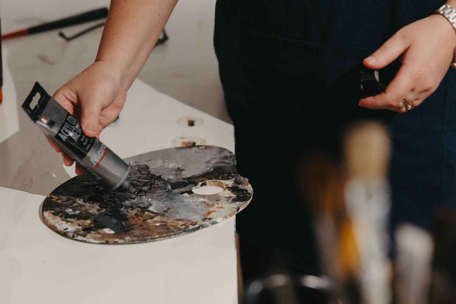 Artist squeezing paint from tube onto wooden palette with brushes in background.