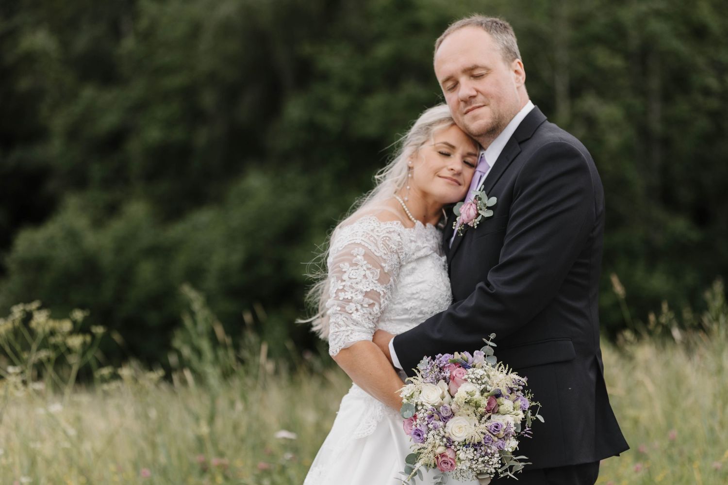 Romantic wedding couple embraces in a sunlit meadow with tall grass and trees in the background.