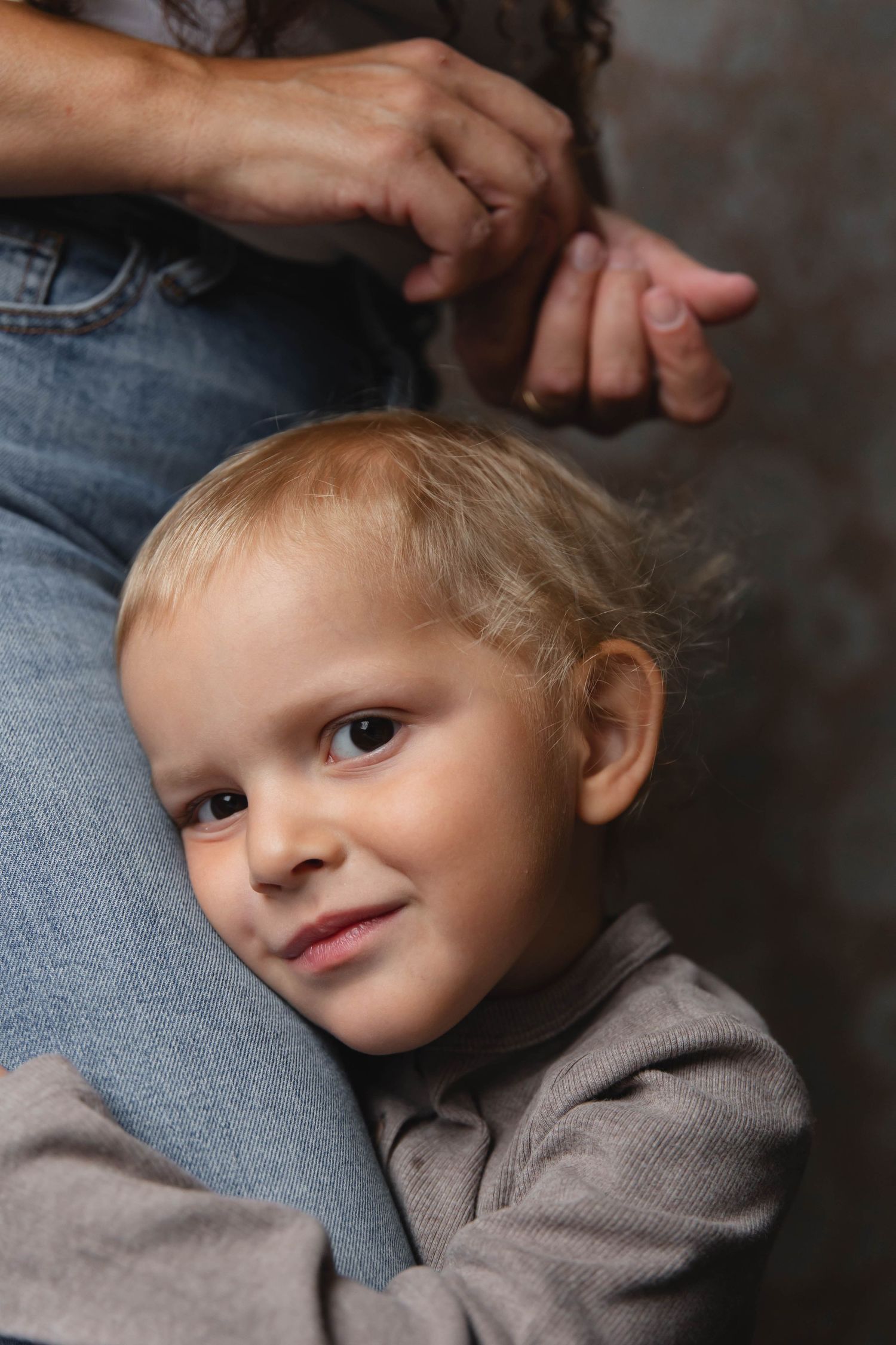 Young child in a gray sweater leaning against someone wearing jeans while smiling softly.