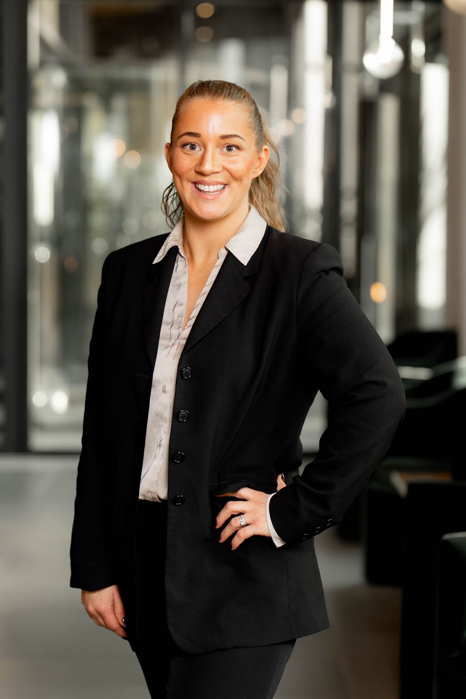 Professional in a black business suit and white blouse stands confidently in an office setting with a bright smile.