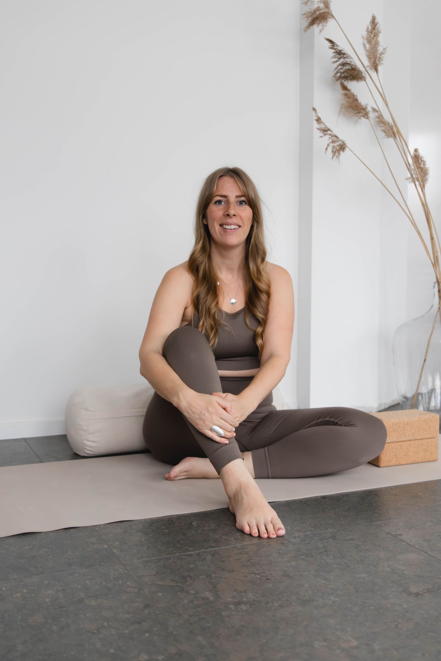 Person in athletic wear sits on floor against white wall with decorative pampas grass in casual lifestyle setting.