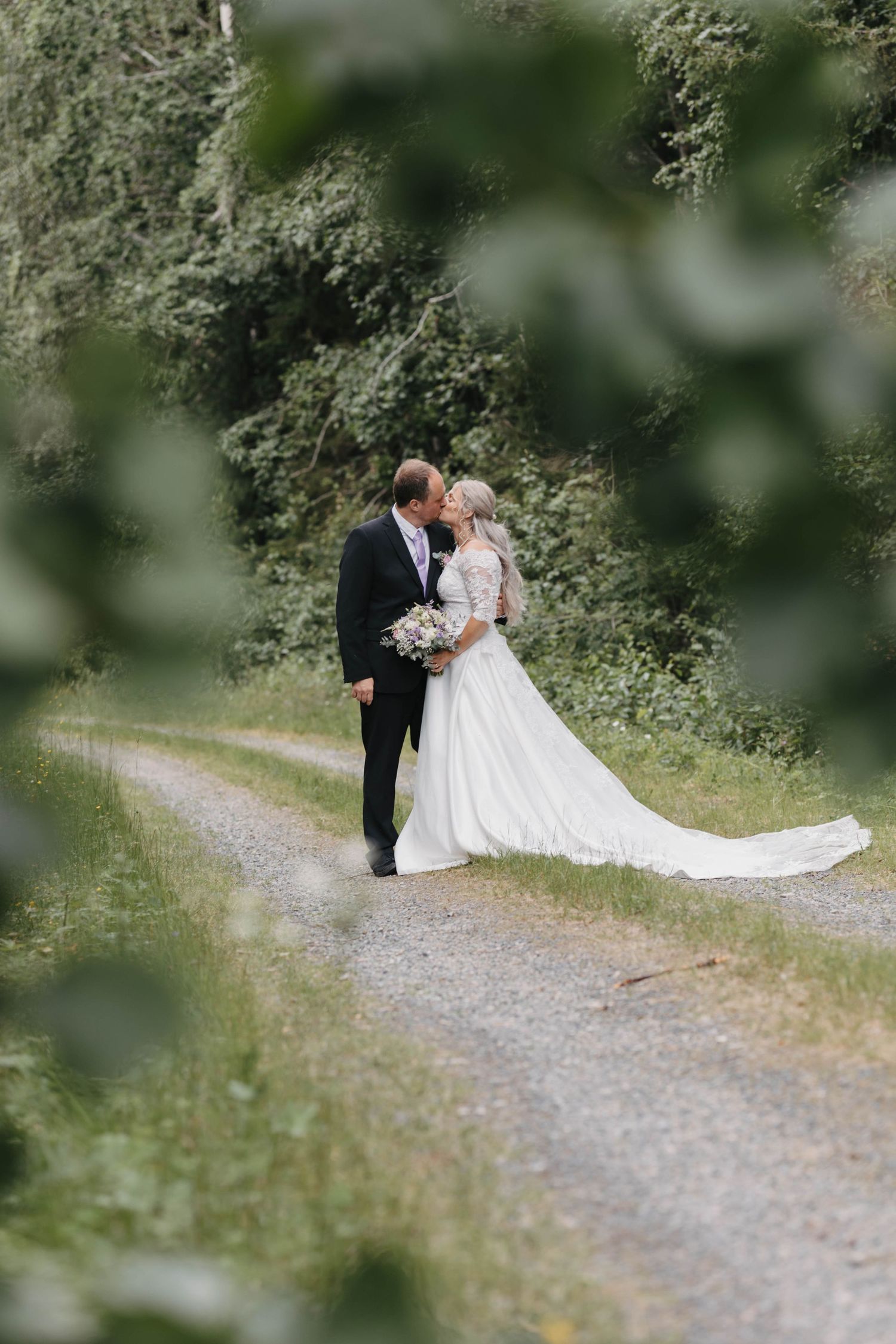A bride and groom share an intimate moment on a gravel path surrounded by lush green foliage.