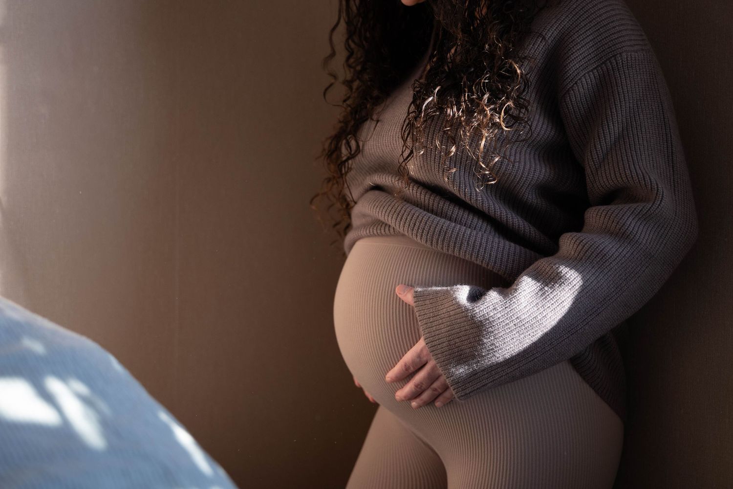 A person wearing a gray sweater sits in a dimly lit room with natural light coming through a window.