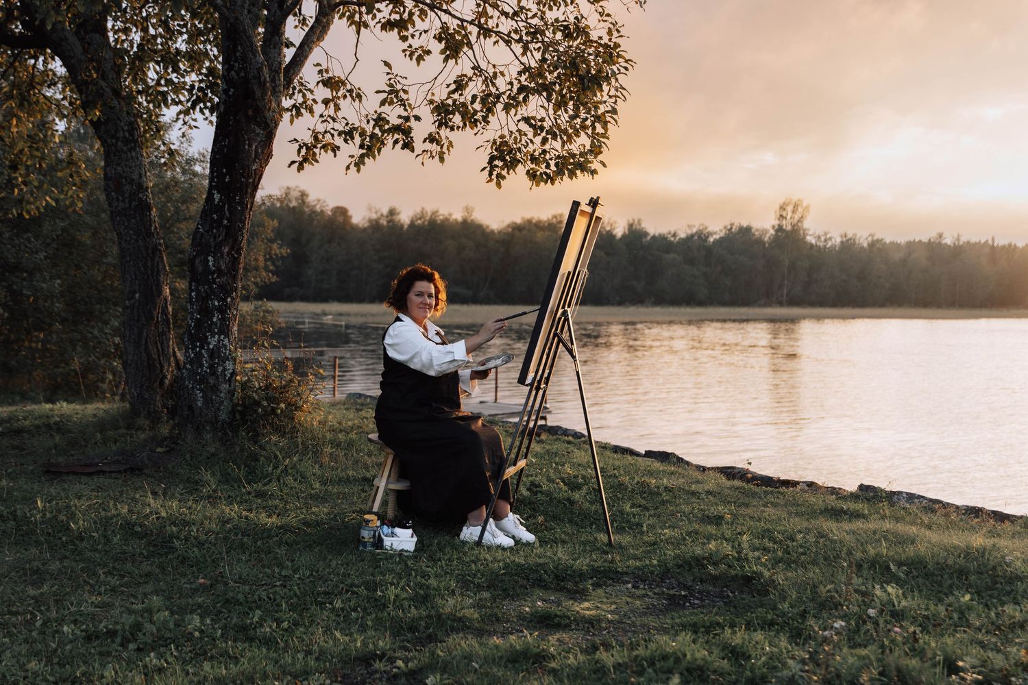 Artist paints on canvas at sunset by tranquil lake with art supplies and easel set up in nature.