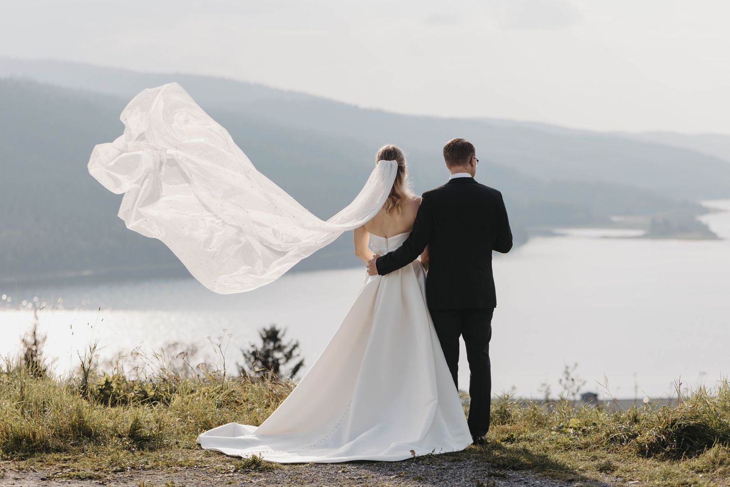 A couple in wedding attire stands on a cliff overlooking a misty lake as the bride's veil flows dramatically in the wind.