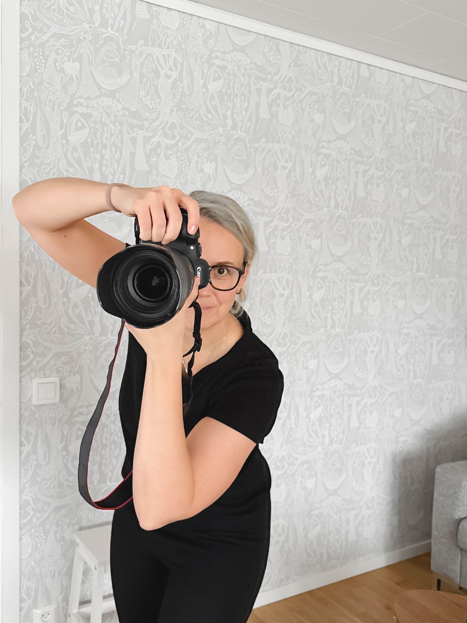 A photographer in a black outfit takes a photo with a professional DSLR camera against a light patterned wallpaper.