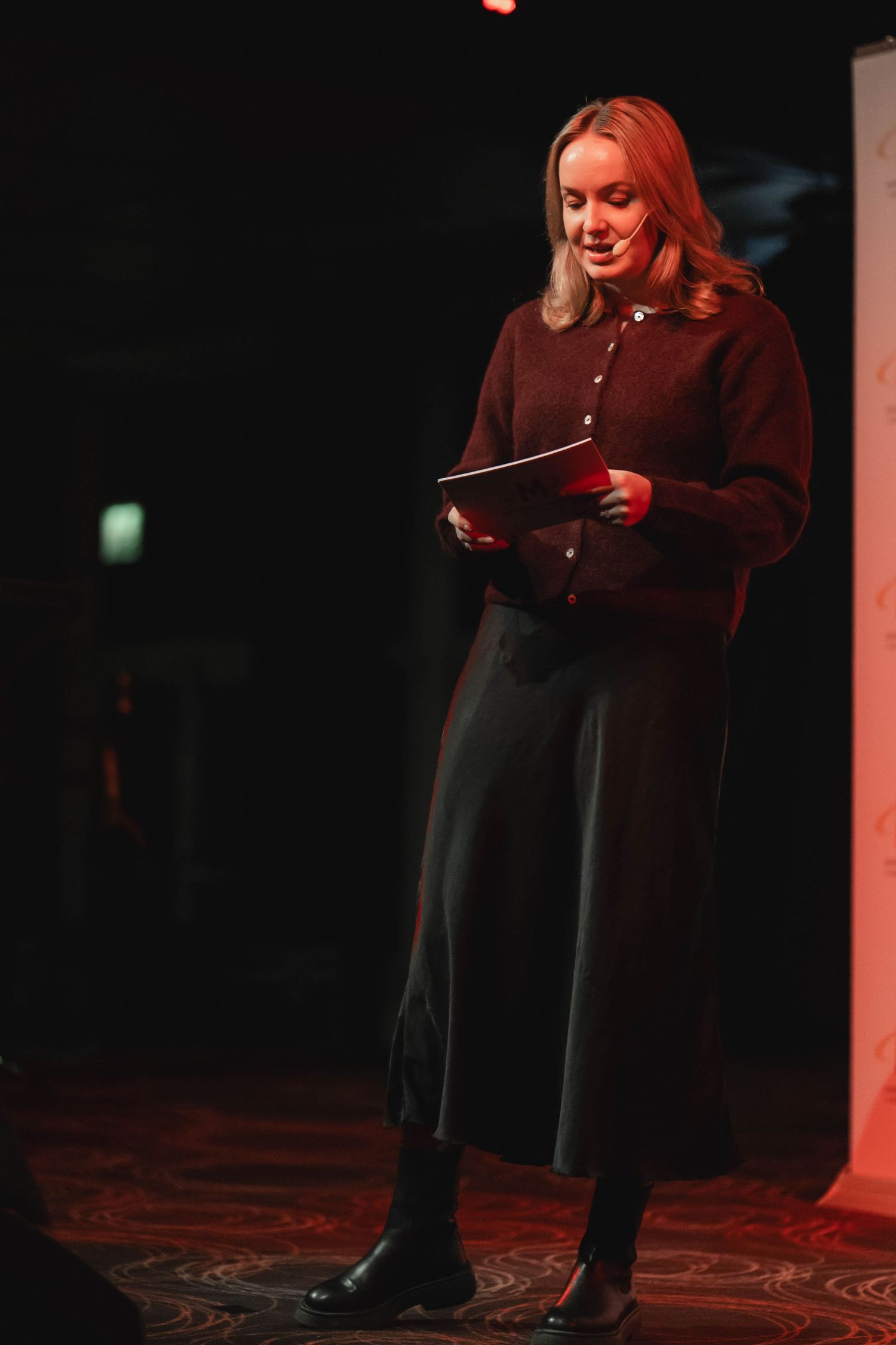A performer in a black dress and brown top stands on stage in dramatic red lighting during a theatrical performance.