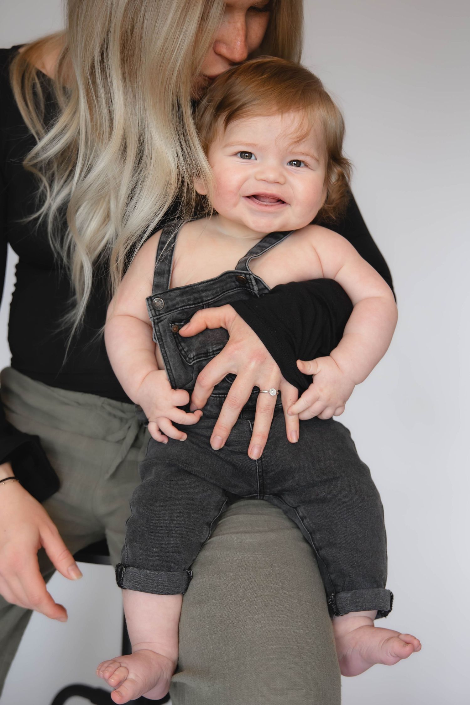 Smiling baby in gray denim overalls being held by a blonde woman in a black top and olive pants.