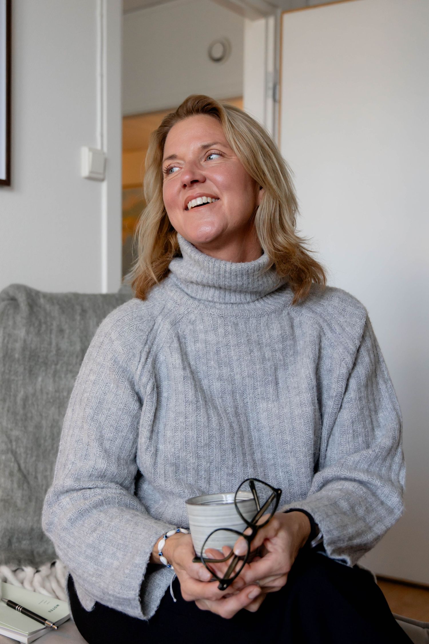 Someone wearing a cozy grey turtleneck sweater sits on a sofa laughing joyfully while holding glasses.