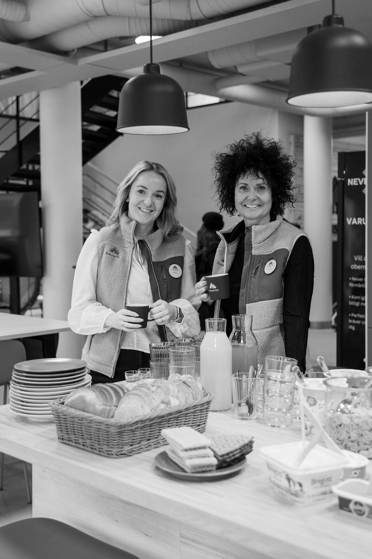 Two people in white uniforms smile while preparing food and drinks at a counter with plates and glassware.