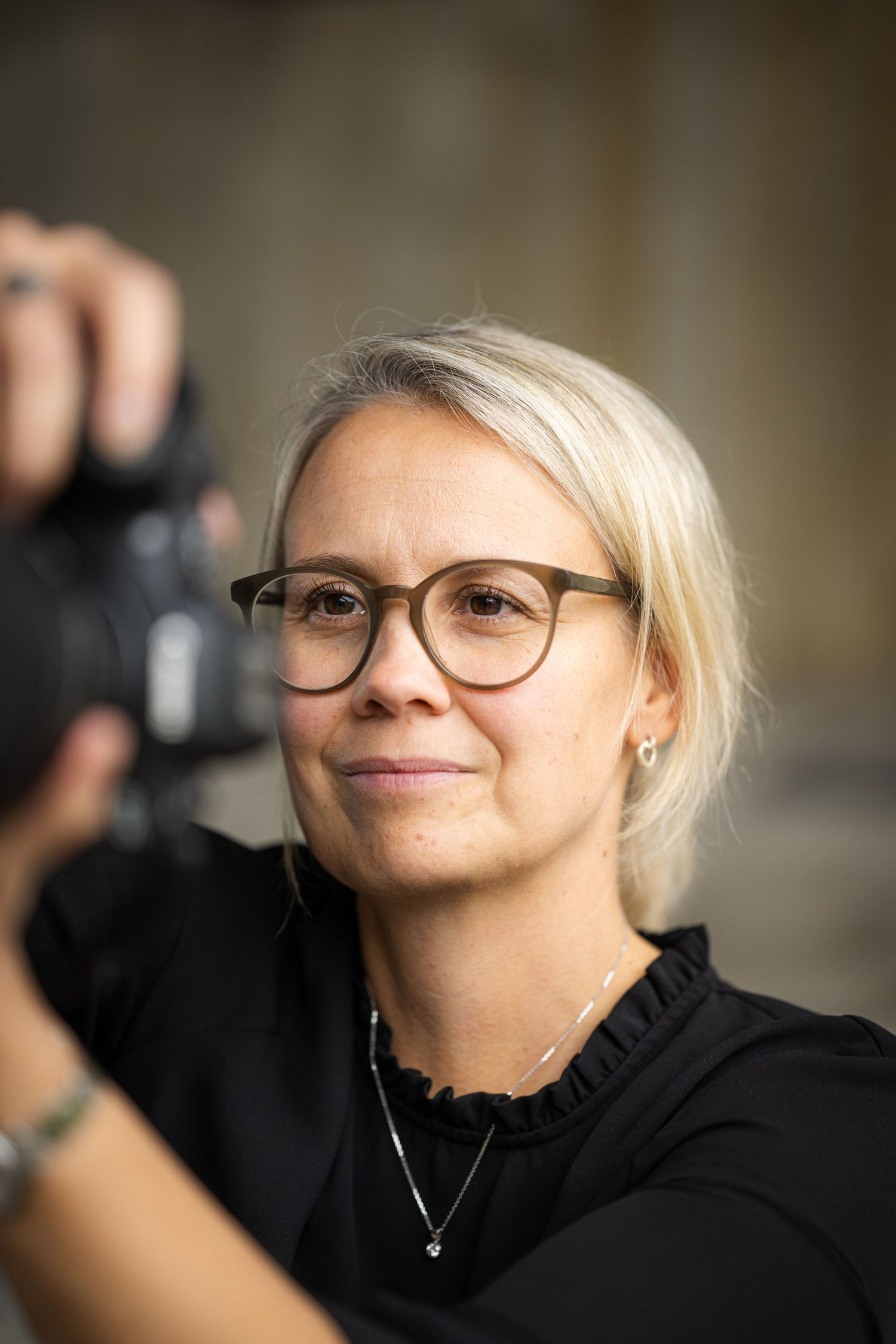 Close up portraits of someone wearing glasses and a black top looking thoughtfully to the side with a slight smile.