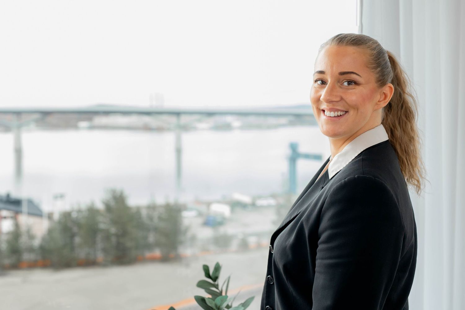 Professional woman in business attire smiles while working at a modern office desk with laptop and plants.