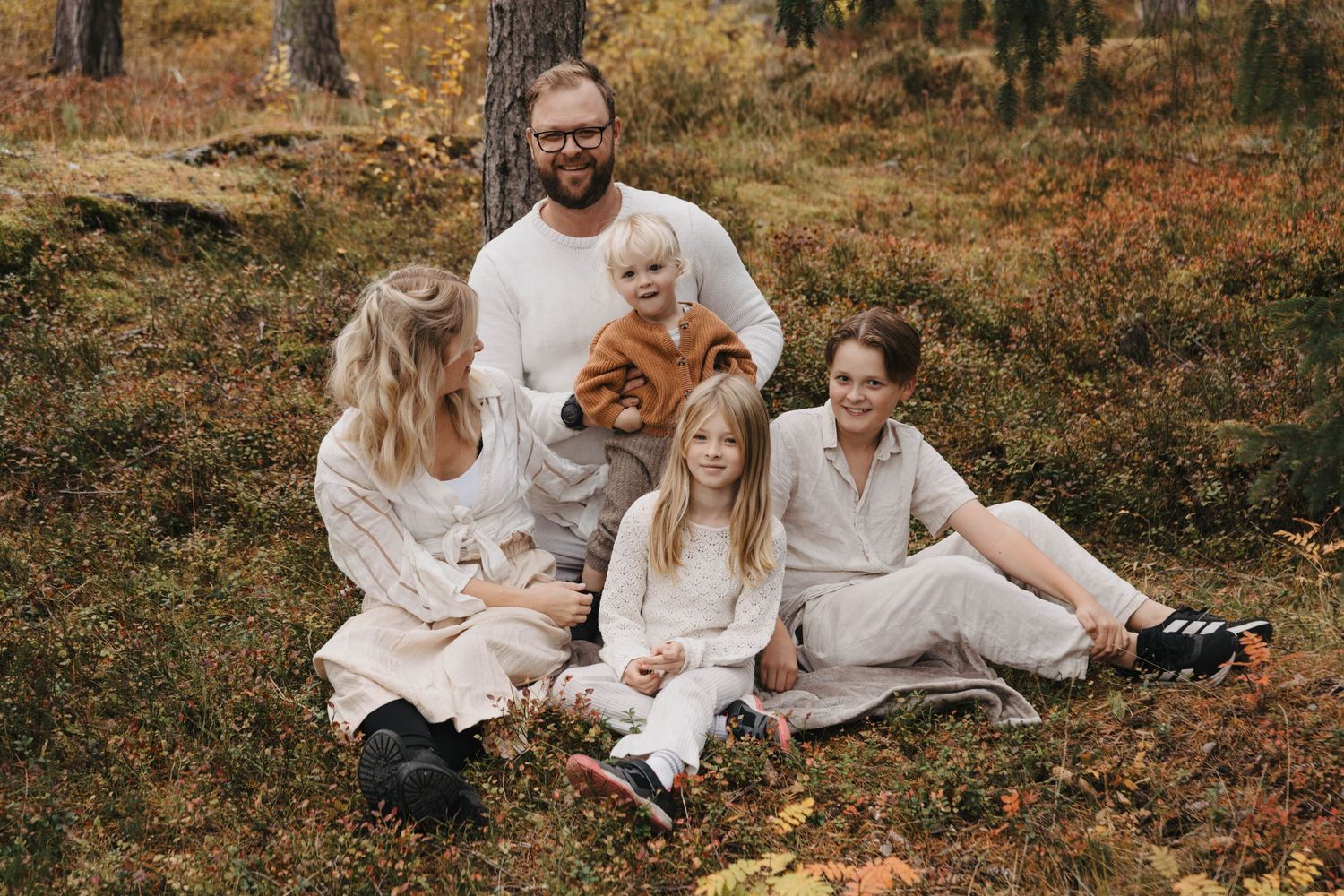 Family wearing white outfits sits together on autumn ground surrounded by fallen leaves in outdoor photo session.