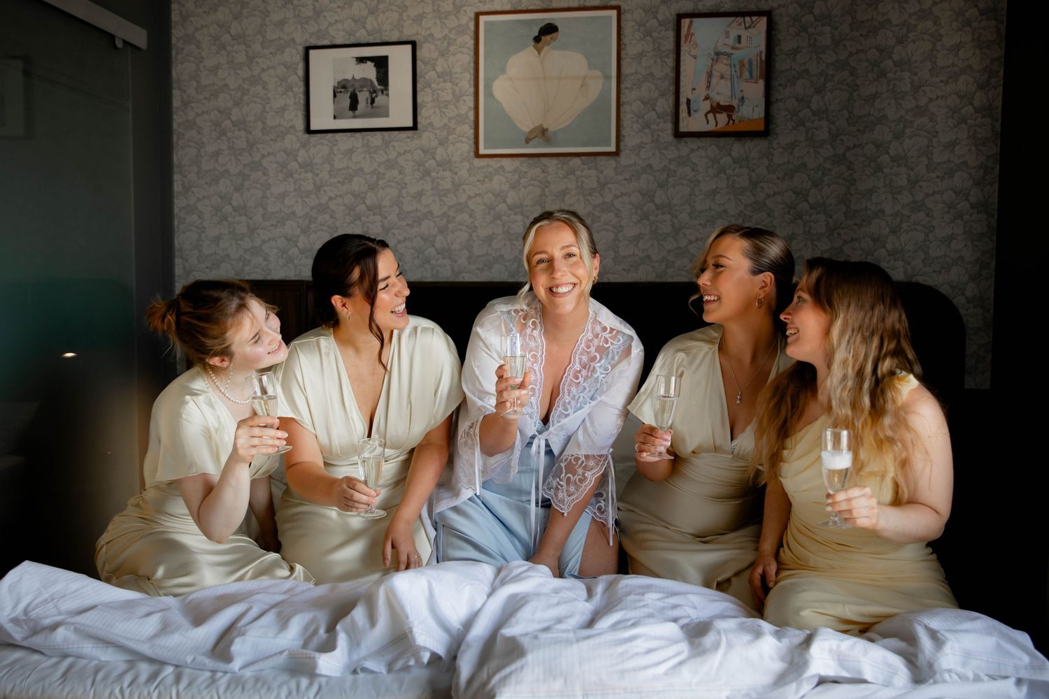 Bride and bridesmaids in silk robes sitting on bed laughing and holding champagne glasses during wedding morning prep.