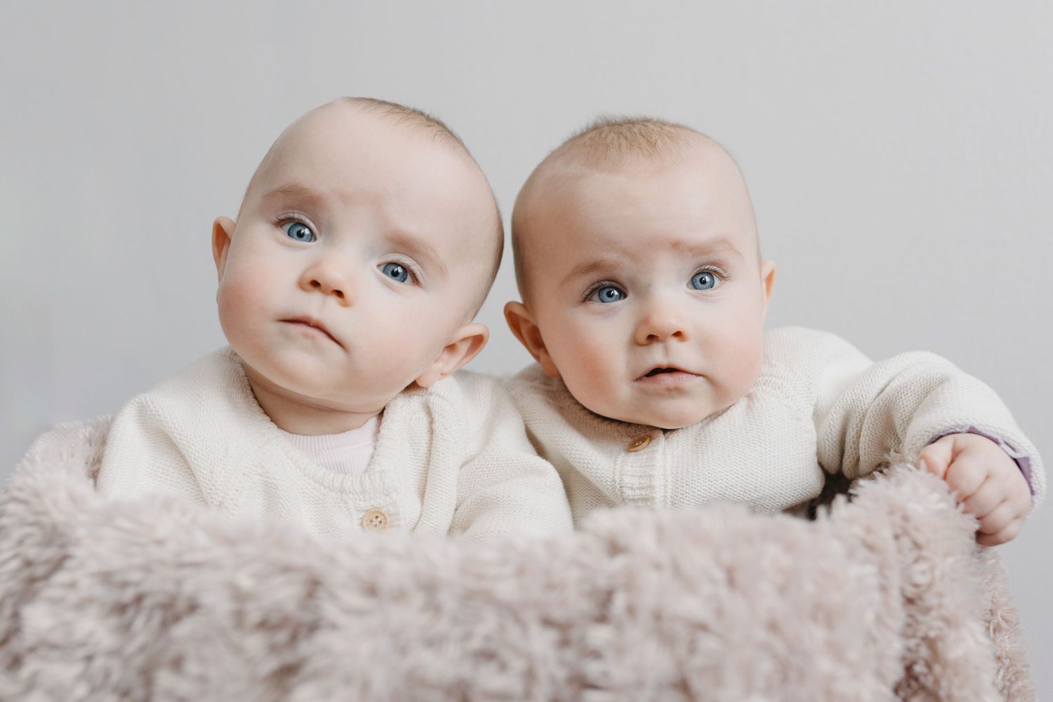 Two adorable babies lying side by side in white outfits against a soft neutral background.