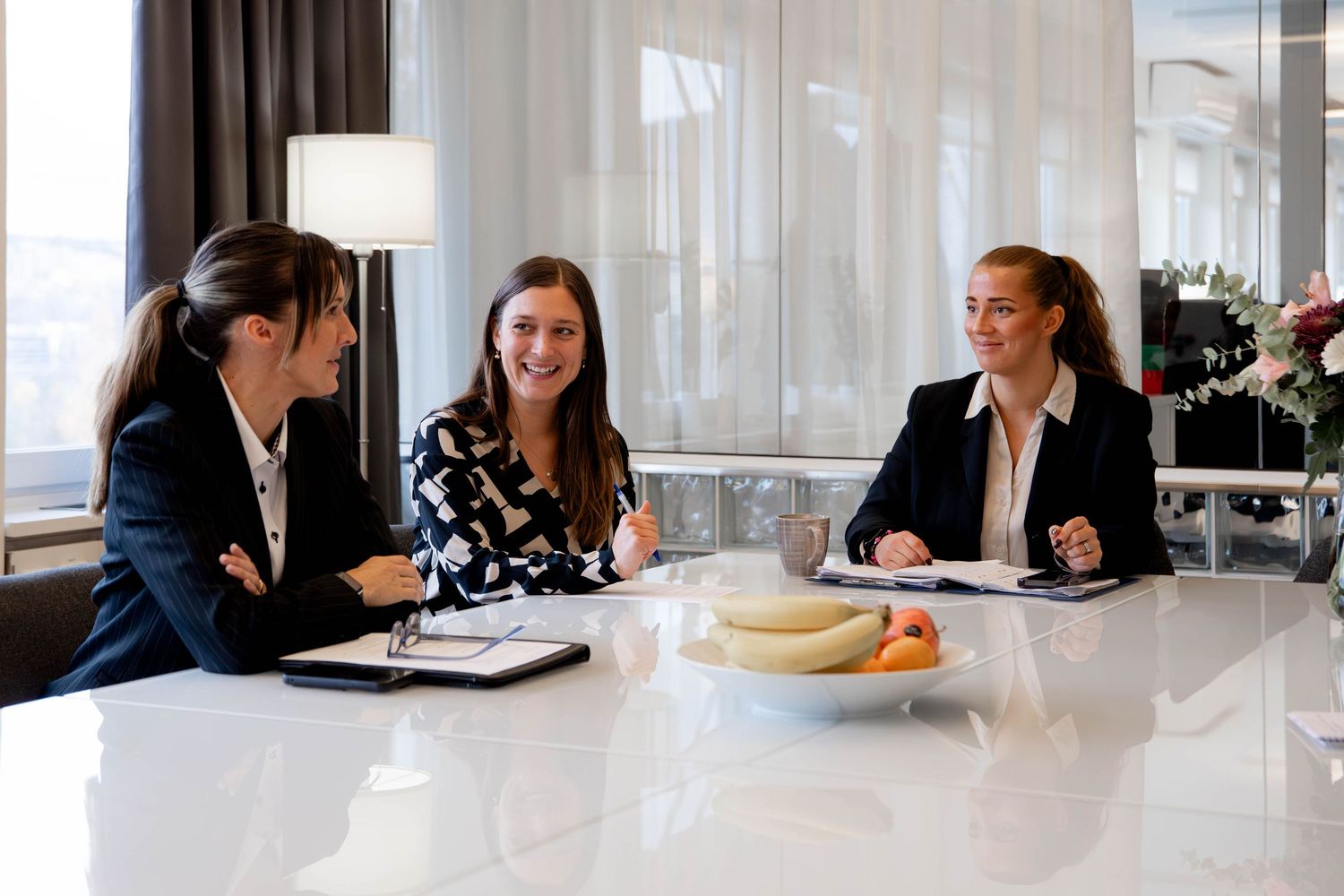 Business professionals sit at a long table during a meeting in a modern office space with natural lighting.
