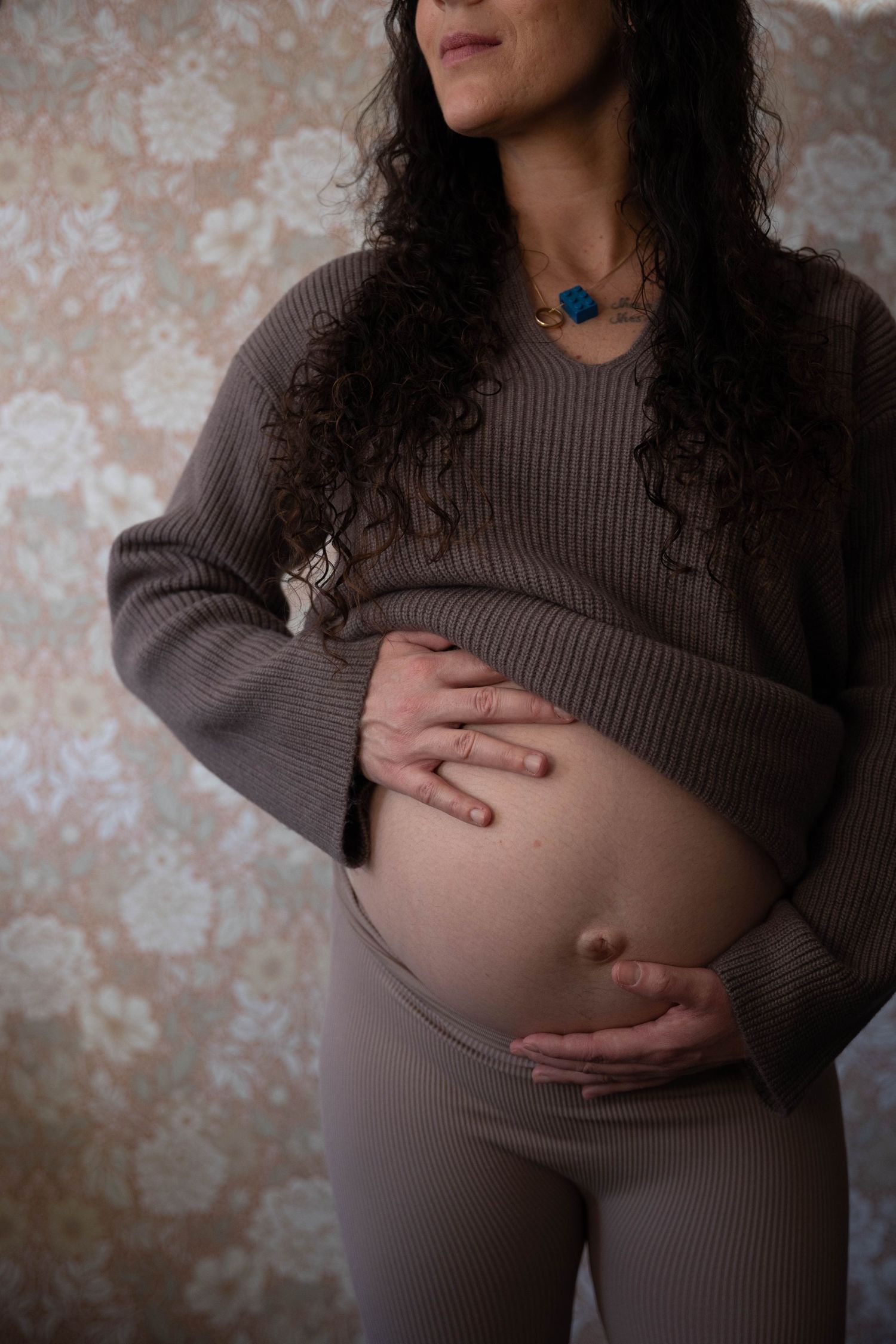 A pregnant woman in a brown sweater gently cradles her baby bump while wearing a blue necklace against a textured wall.