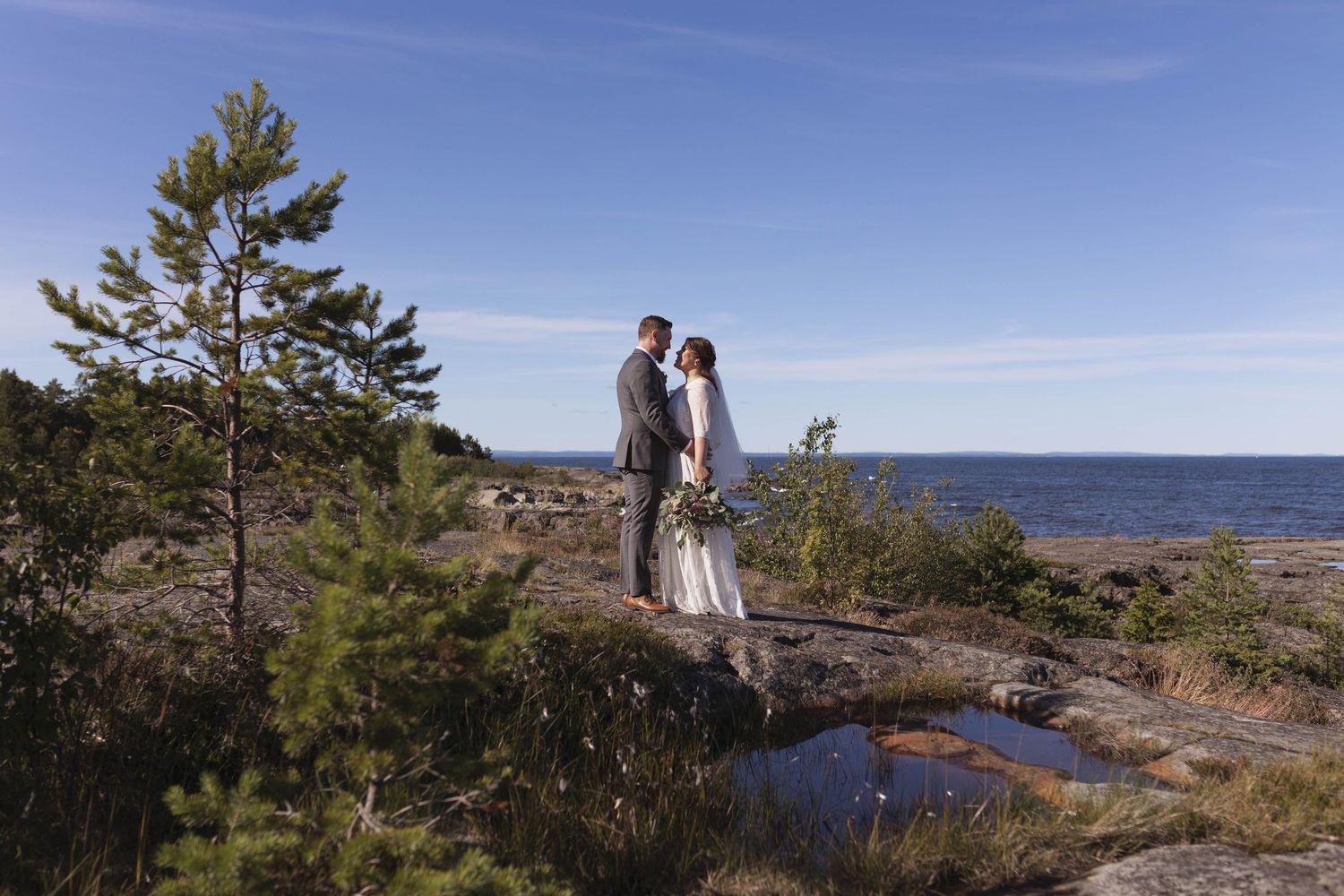 A coastal wedding ceremony takes place on a rocky cliff overlooking the ocean on a sunny day.