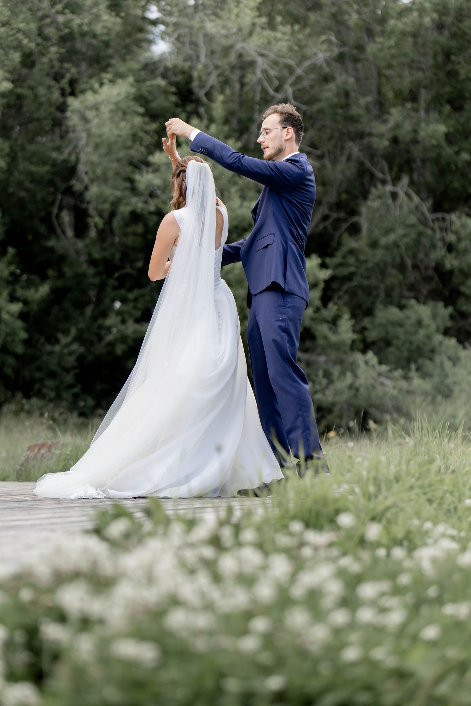 Groom in navy suit twirling bride in white wedding gown and veil on wooden path surrounded by greenery.