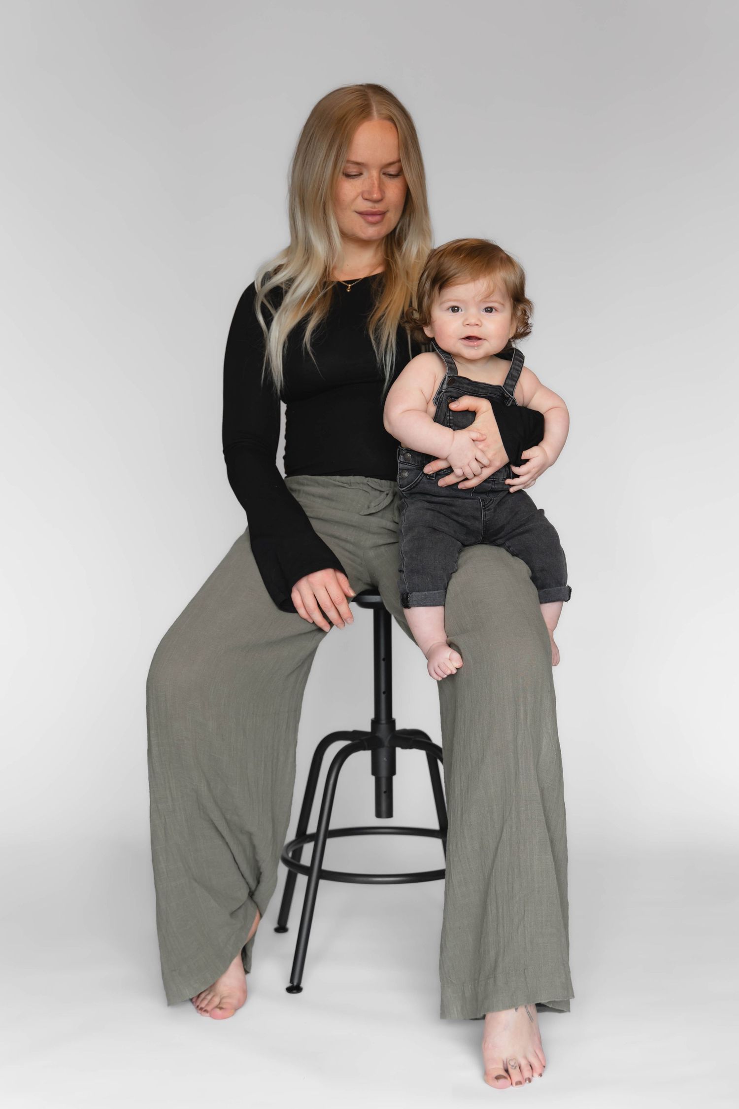 Woman in black top and olive wide-leg pants sits on stool holding a baby in dark overalls.