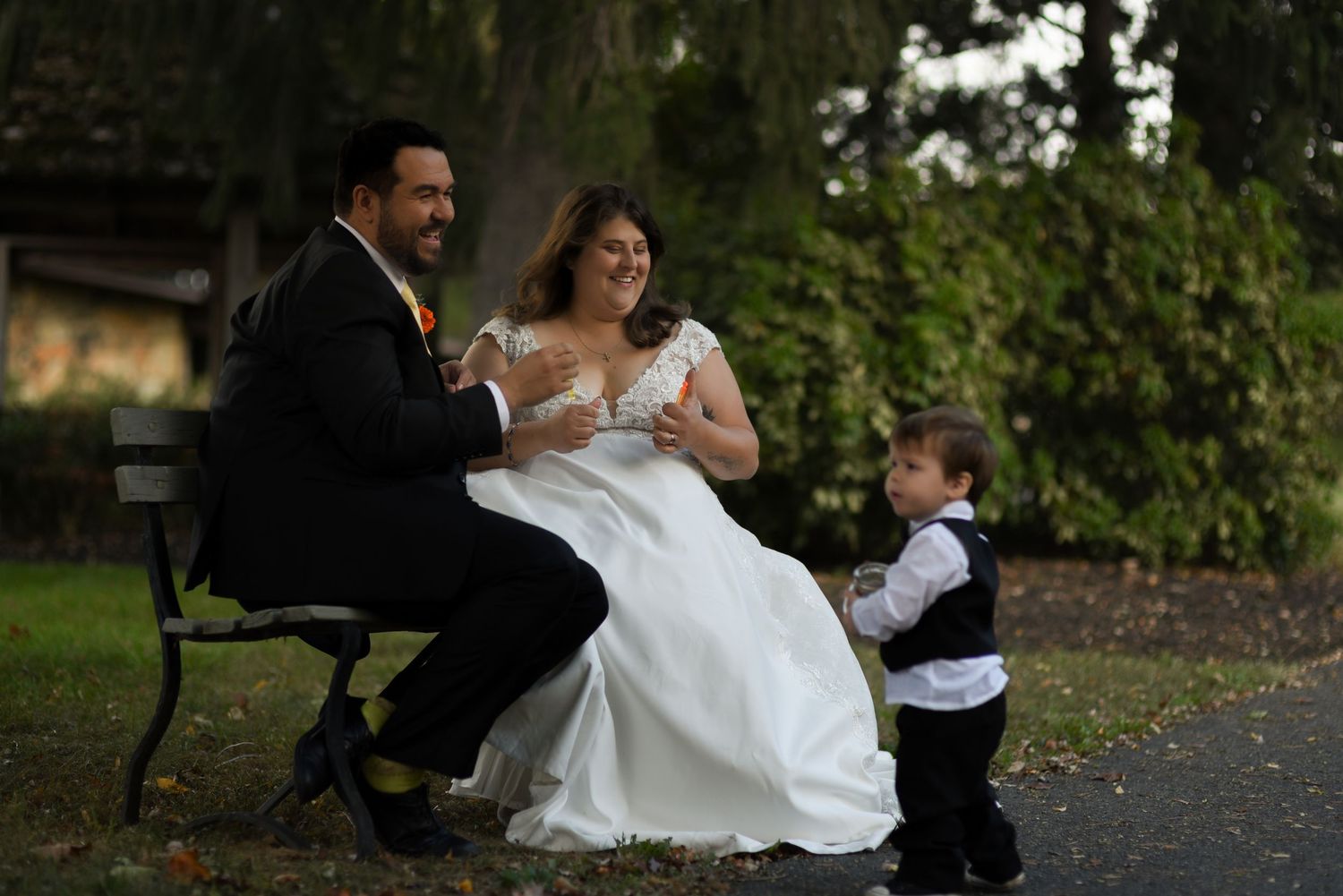 Groom and bride right after blowing bubbles, sitting on a park bench outside the reception with their young son