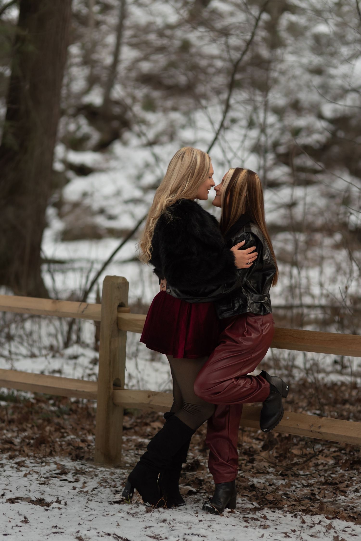 Wives going in for a kiss while leaning against a wooden fence with a snowy creek behind them