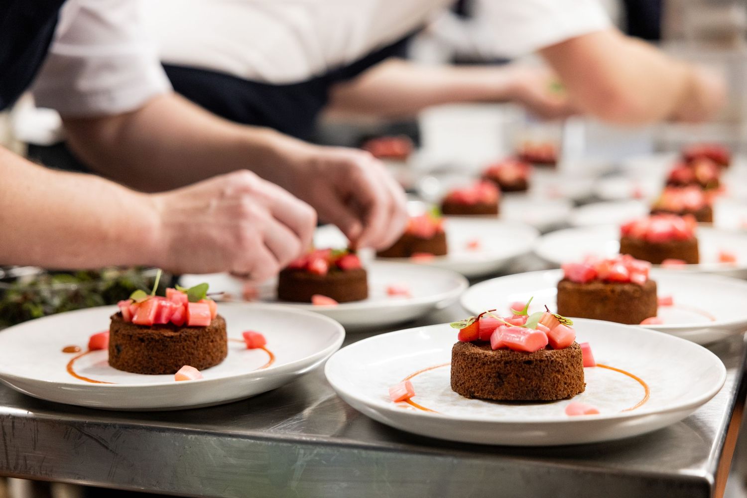 Chef plating multiple identical desserts on white plates in restaurant kitchen.