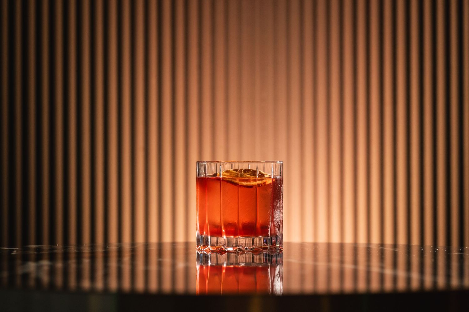 Amber-colored cocktail in crystal glass against striped background with dramatic lighting.