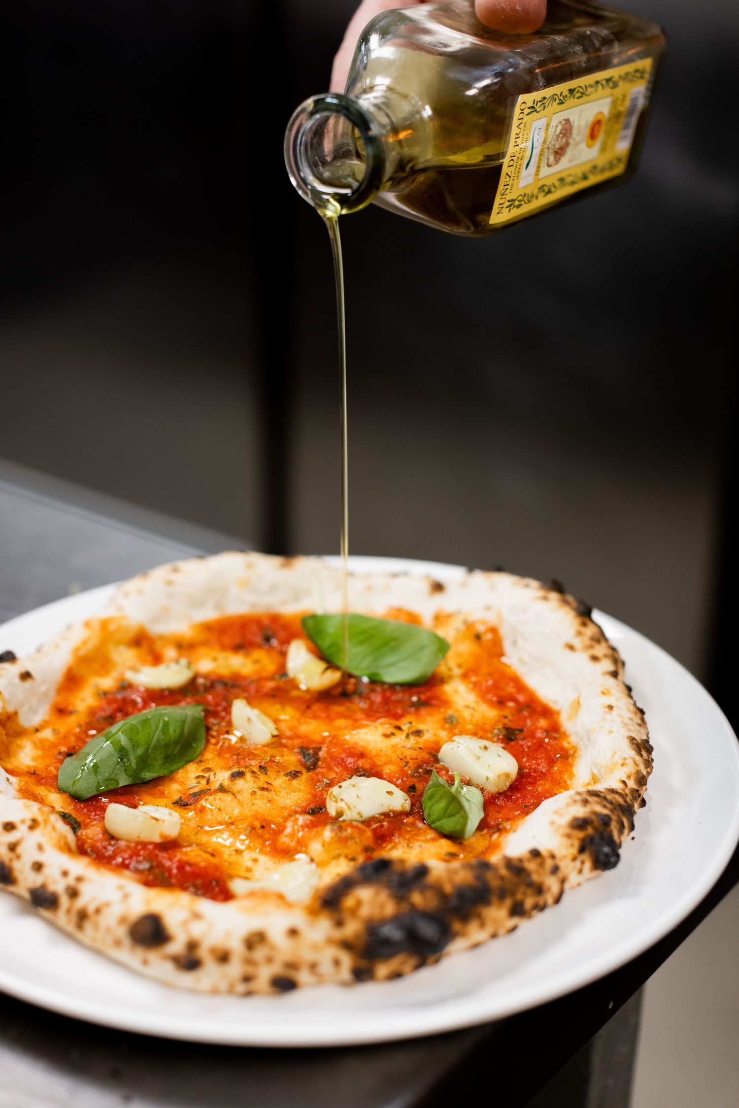 Olive oil being drizzled over a Neapolitan Margherita pizza with fresh basil leaves.
