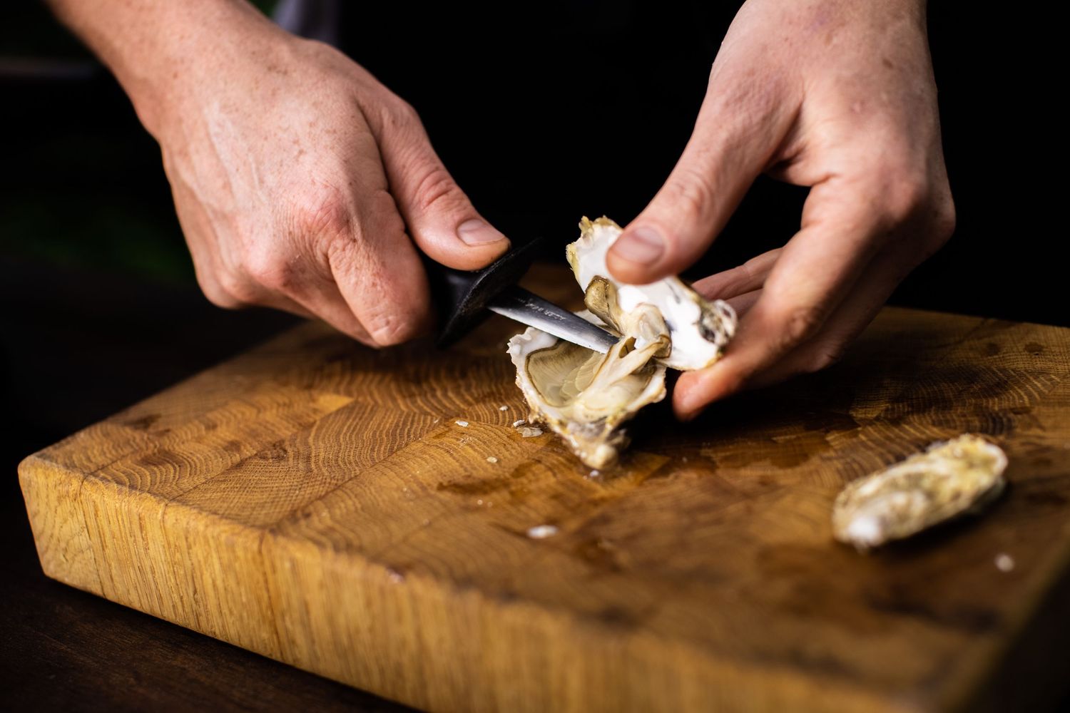 Hands slicing fresh bread on wooden cutting board in dark setting.