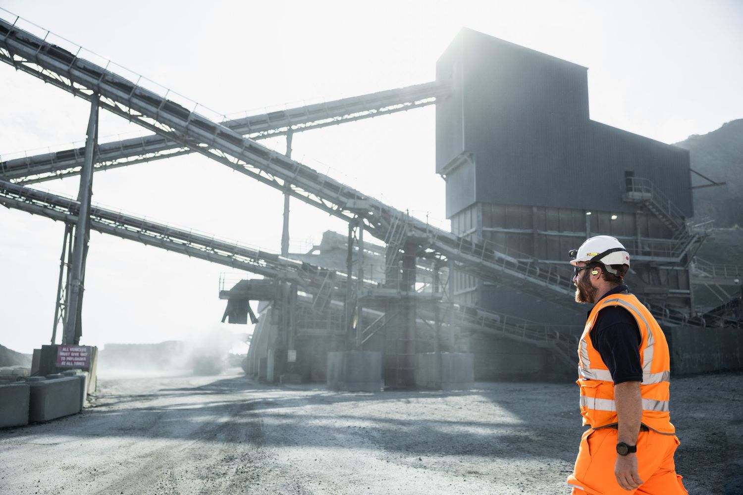 Worker in high visibility gear observing industrial facility with conveyor systems.