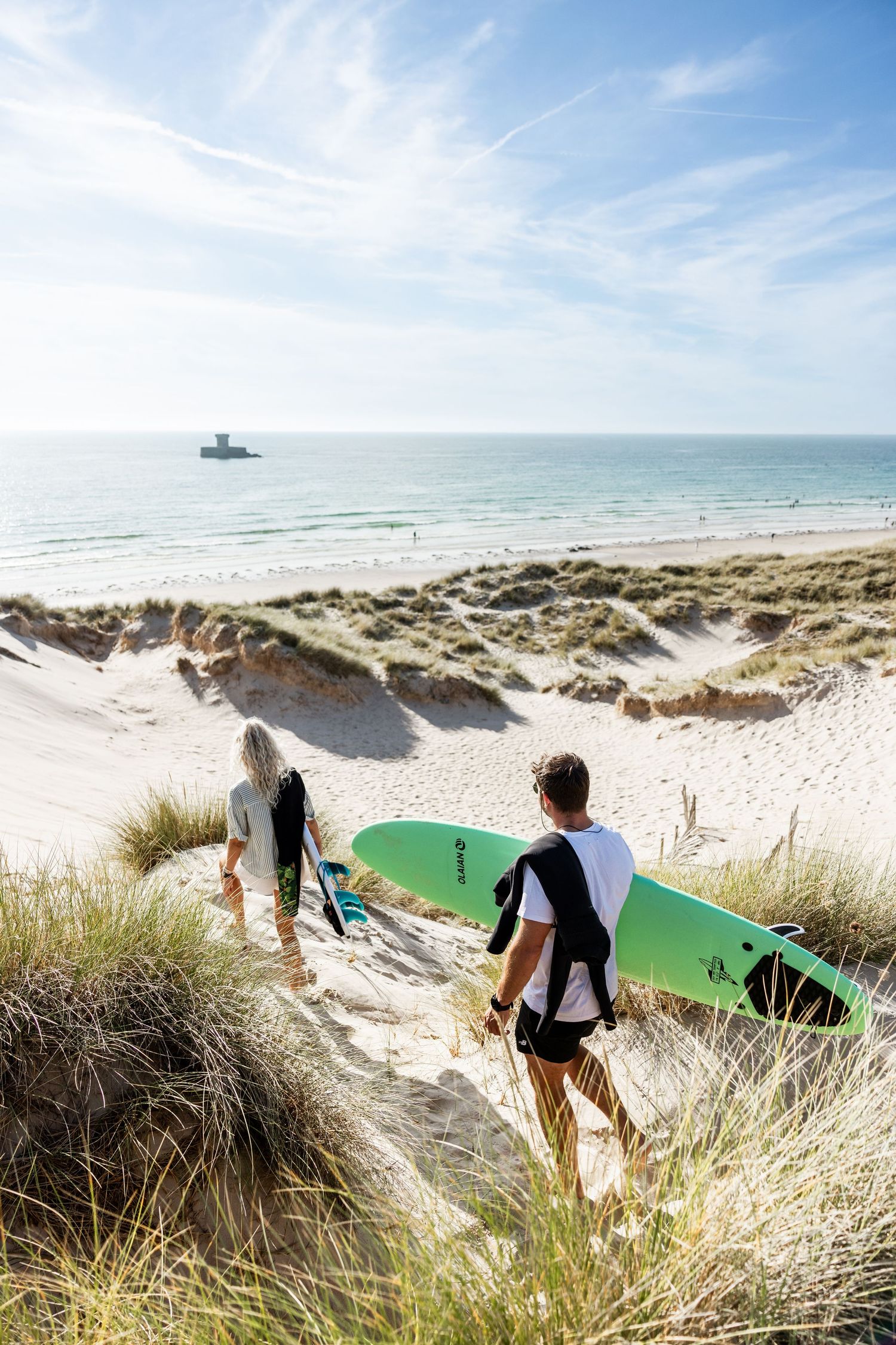 Surfers carrying colorful boards down grass-covered sand dunes towards the beach.