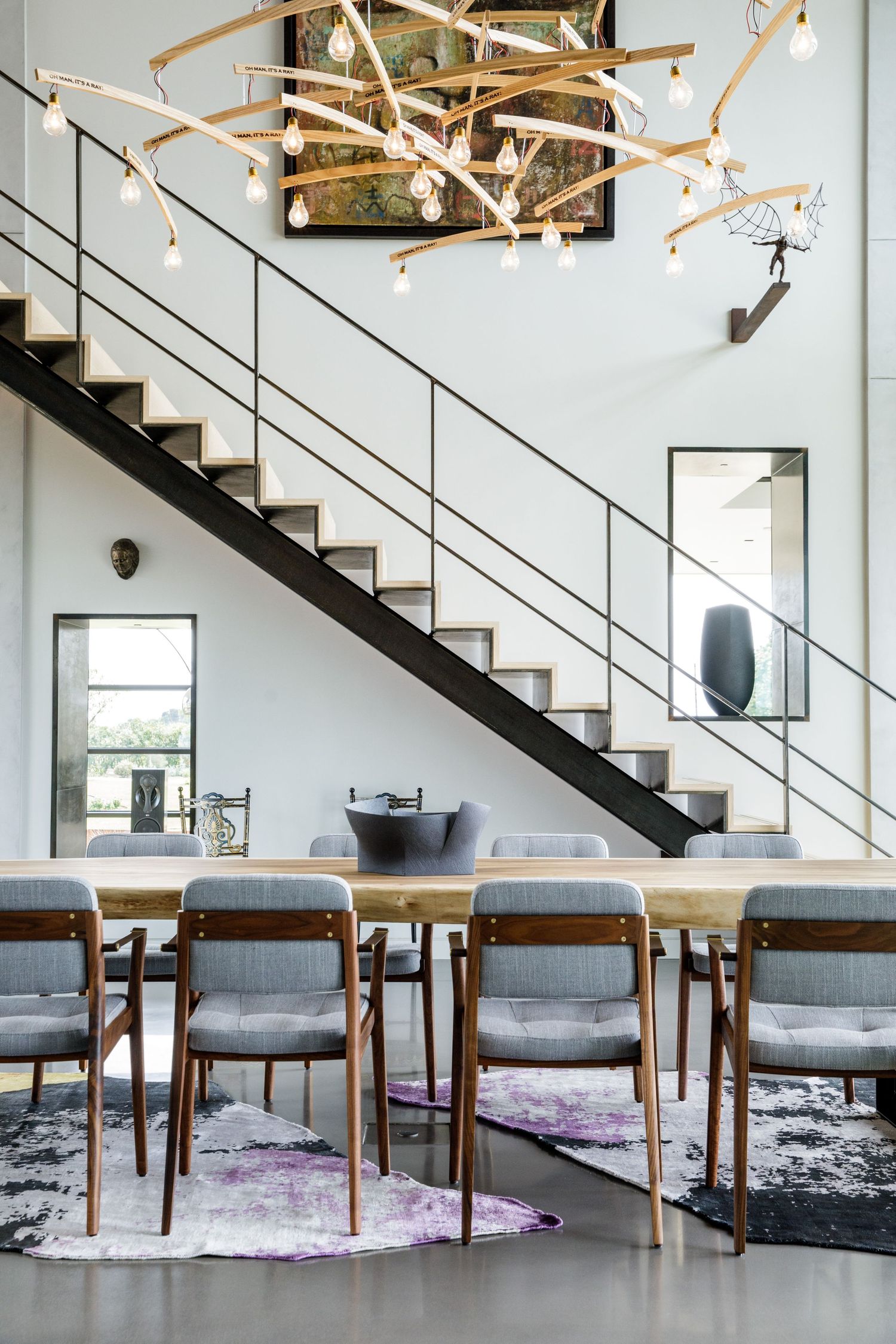 Modern dining area with grey upholstered chairs and wooden table under geometric light fixtures and black staircase.