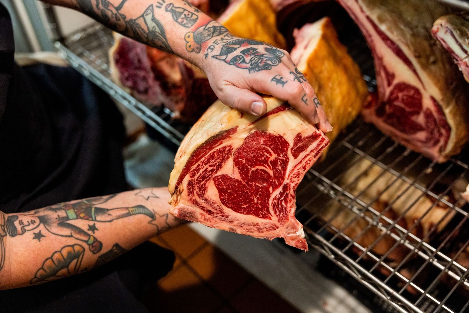 Tattooed hands holding premium cuts of raw meat in a restaurant kitchen.