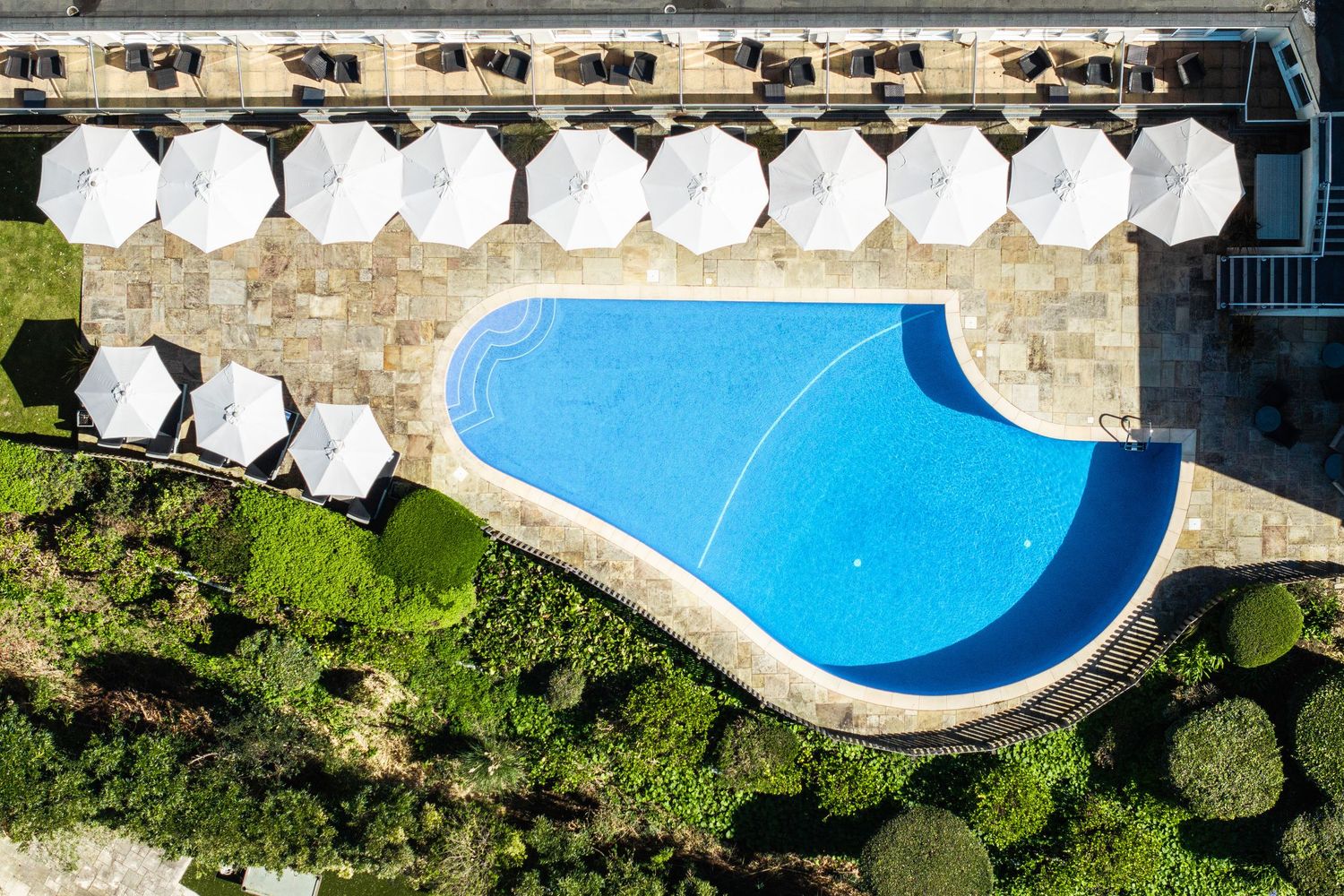 Aerial view of a luxury hotel swimming pool surrounded by white umbrellas and stone architecture.