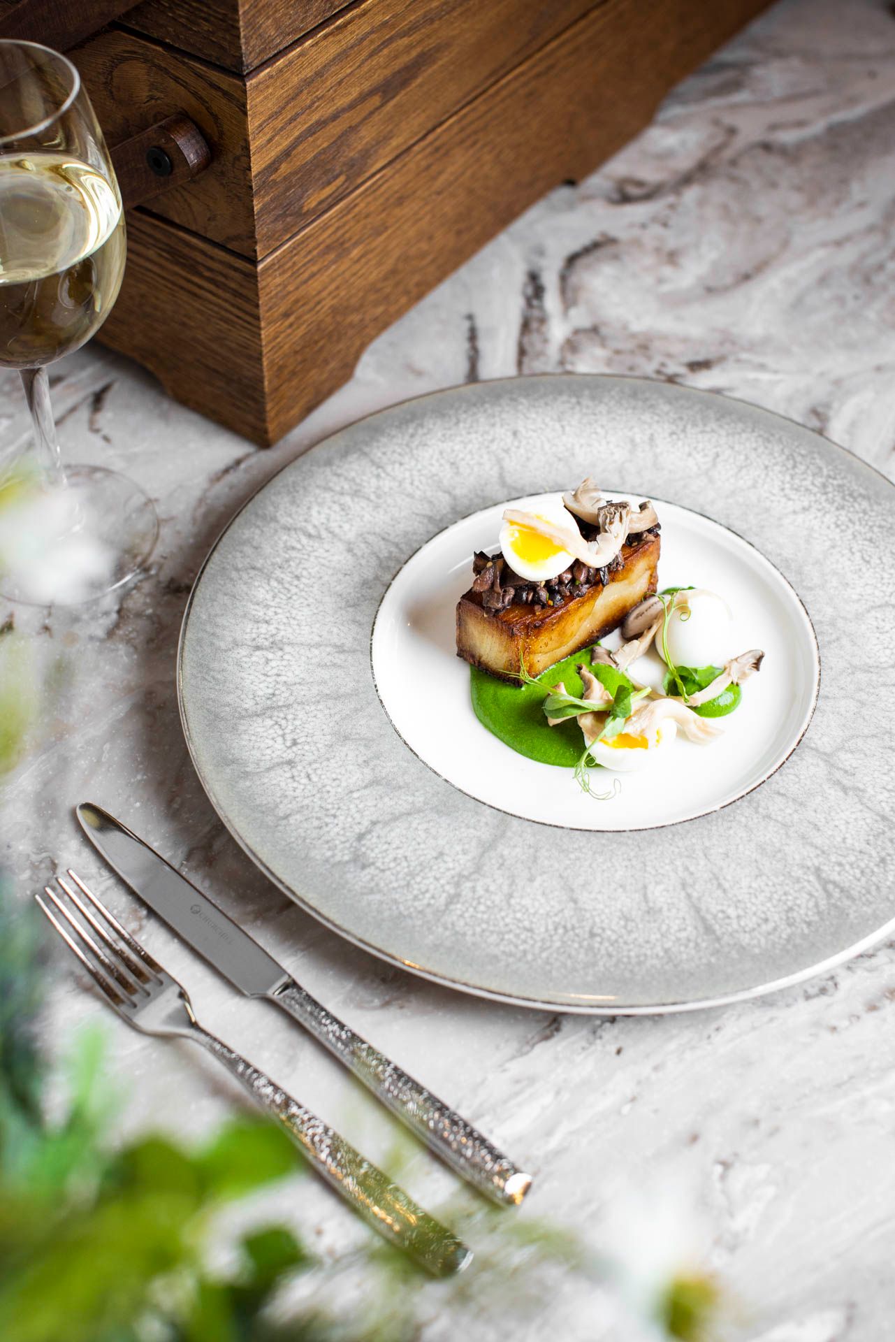Elegant plated dish with quail egg and microgreens on textured white plate beside wine glass.
