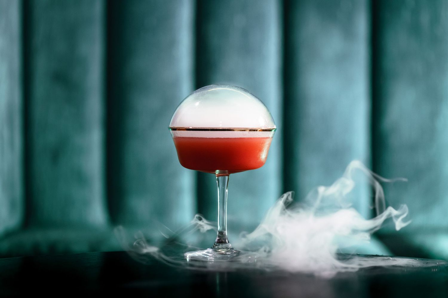 Smoke swirls around red cocktail in spherical glass against teal backdrop.