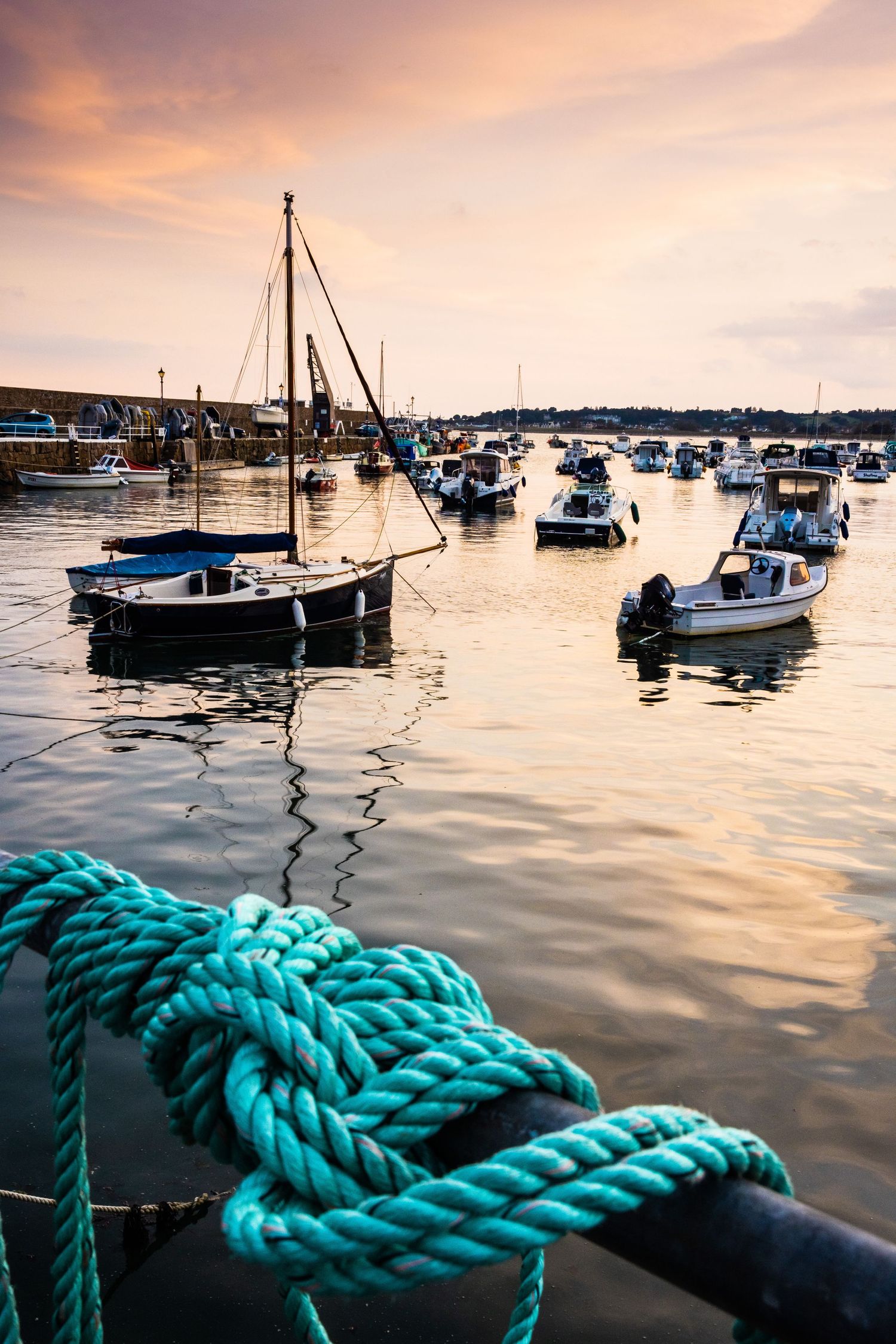 Turquoise mooring rope tied on dock post with boats anchored in harbor during sunset.
