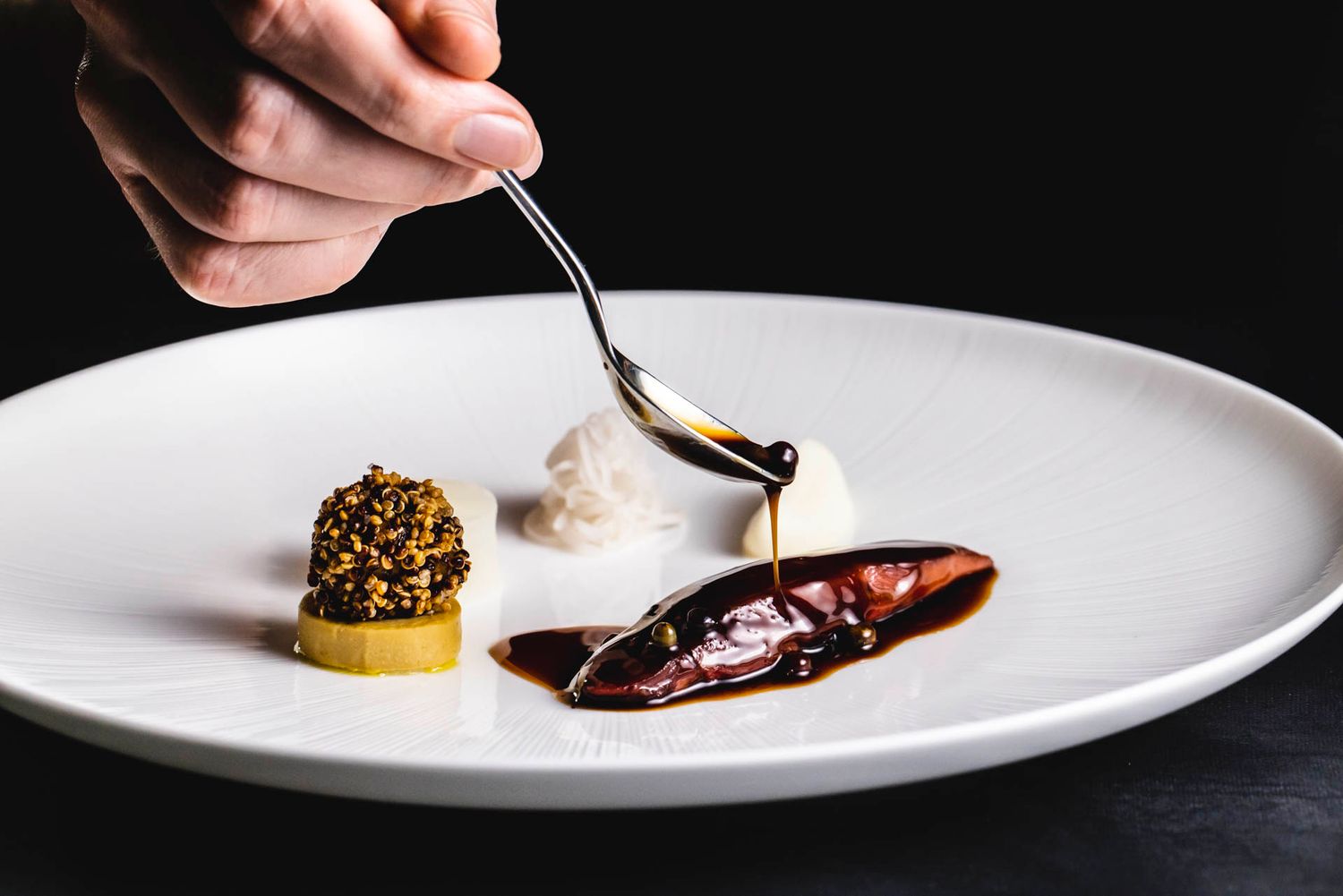 Fine dining plating with a sauce being drizzled over an elegantly presented dish on a white plate.