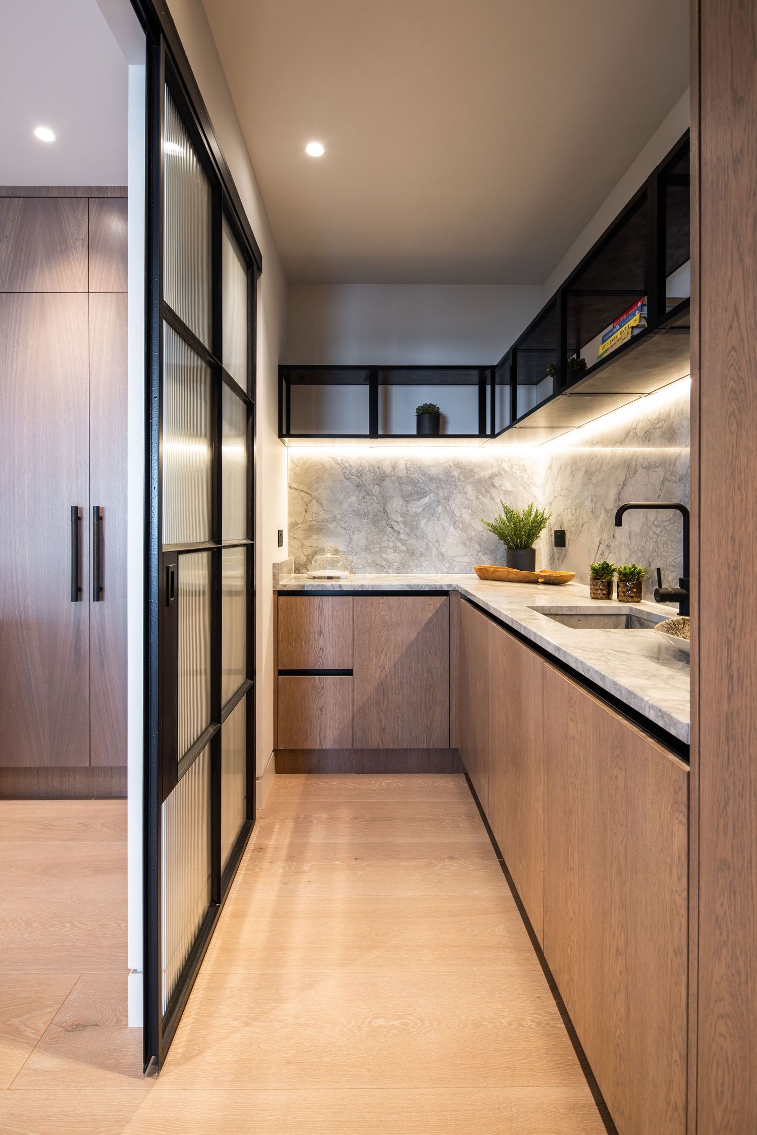 Modern galley kitchen with wood cabinets, marble backsplash, and black-framed glass doors.